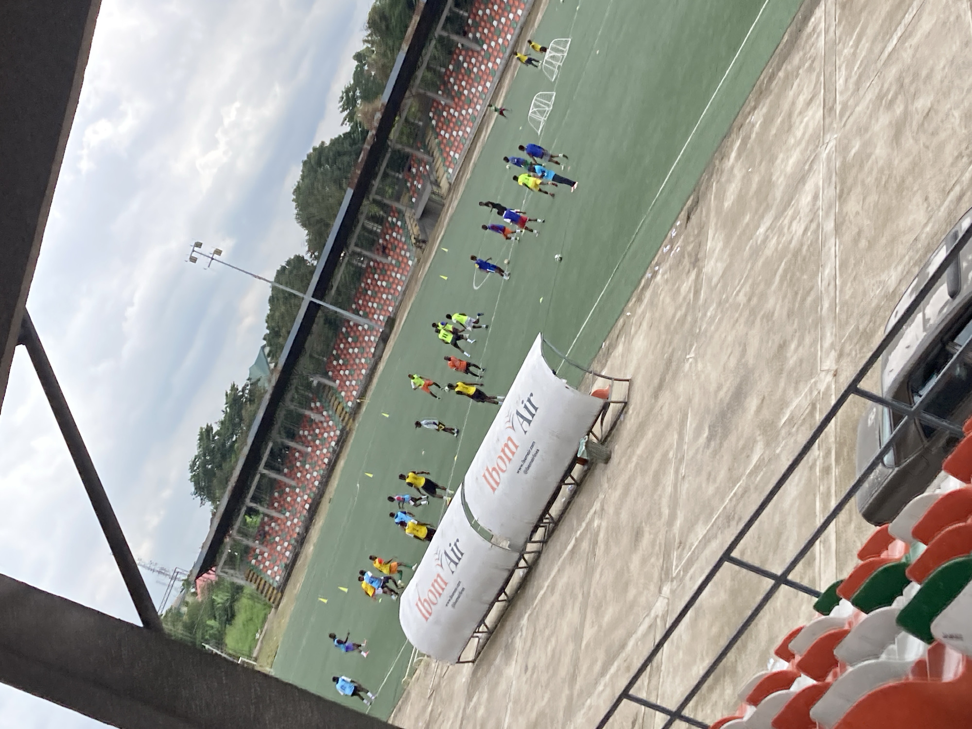 The image shows a training session of a soccer team on a green artificial turf field, likely at the Godswill Akpabio International Stadium in Uyo, Nigeria, due to the "Ibom Air" branding visible on the dugout. Multiple male players, wearing various colored jerseys and shorts, are spread across the field, some engaging with soccer balls. Several players appear to be dribbling or passing, indicating active practice. The field is marked with white lines and dotted with yellow cones and small portable goals, suggesting drills are in progress. In the background, tiered stadium seating with alternating red, white, and green seats is visible. A covered dugout, also branded with "Ibom Air," is positioned near the sideline. Beyond the stadium, trees and buildings can be seen, and the sky is overcast with scattered clouds. A dark colored car is parked in the lower right corner, near the seating area.