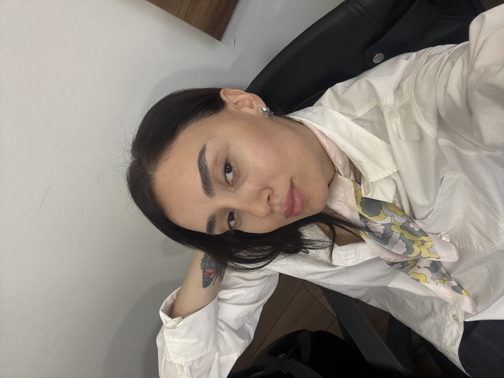 A woman with dark hair and a butterfly tattoo is seated. She is wearing a white shirt and a patterned scarf. She appears to be in an indoor setting, possibly an office or home. The location is unknown.