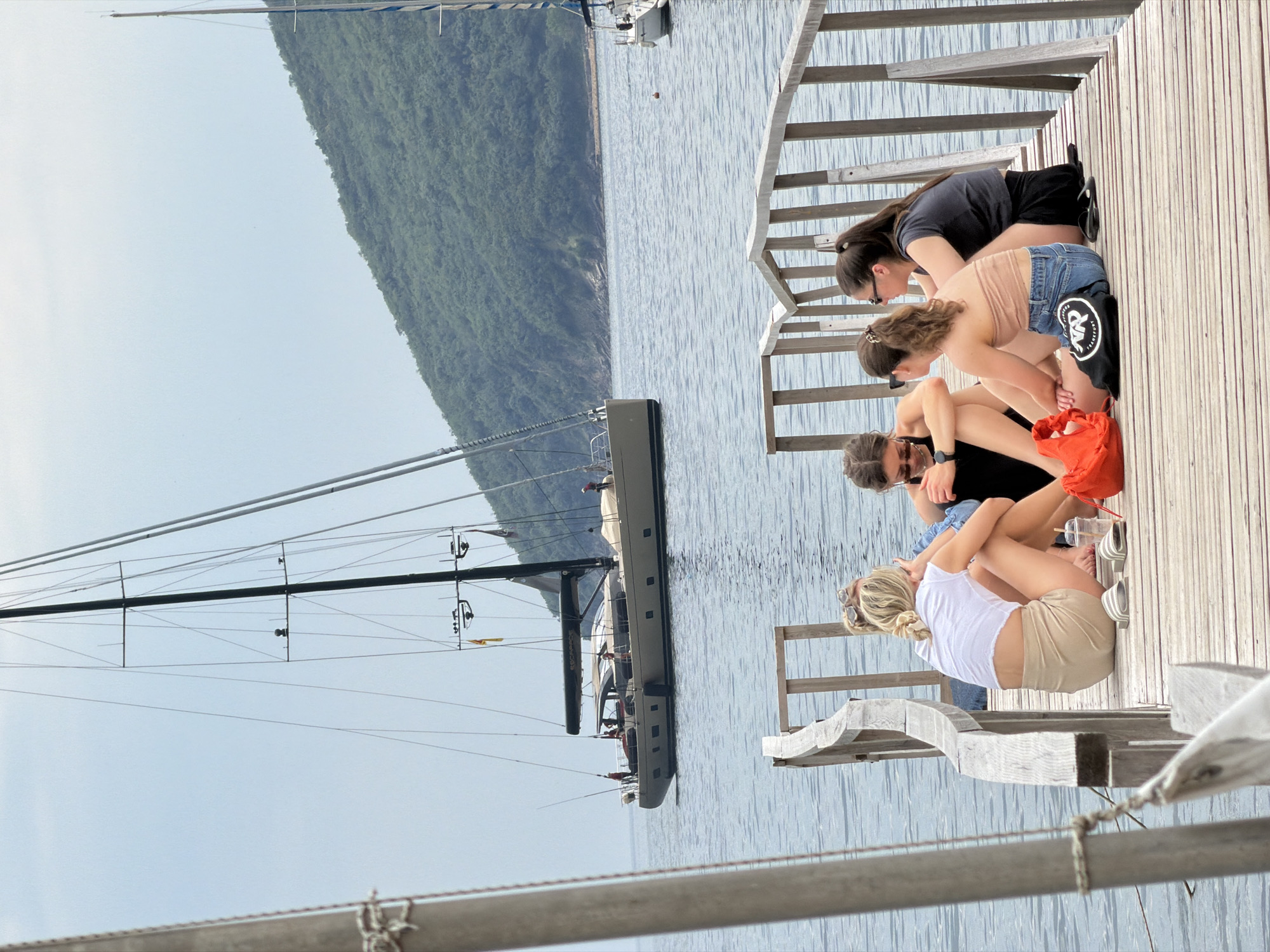 Four young women are sitting on a wooden dock in Budva, Montenegro. The dock extends out into the Adriatic Sea, with a large sailboat anchored nearby. A lush green mountain range is visible across the bay. The women are dressed casually in shorts, t-shirts, and tank tops. One of them is wearing sunglasses and a watch. An orange bag and a pair of sandals are resting on the dock. The scene is sunlit and the water is calm. The wooden dock and guard rails are built of weathered planks.