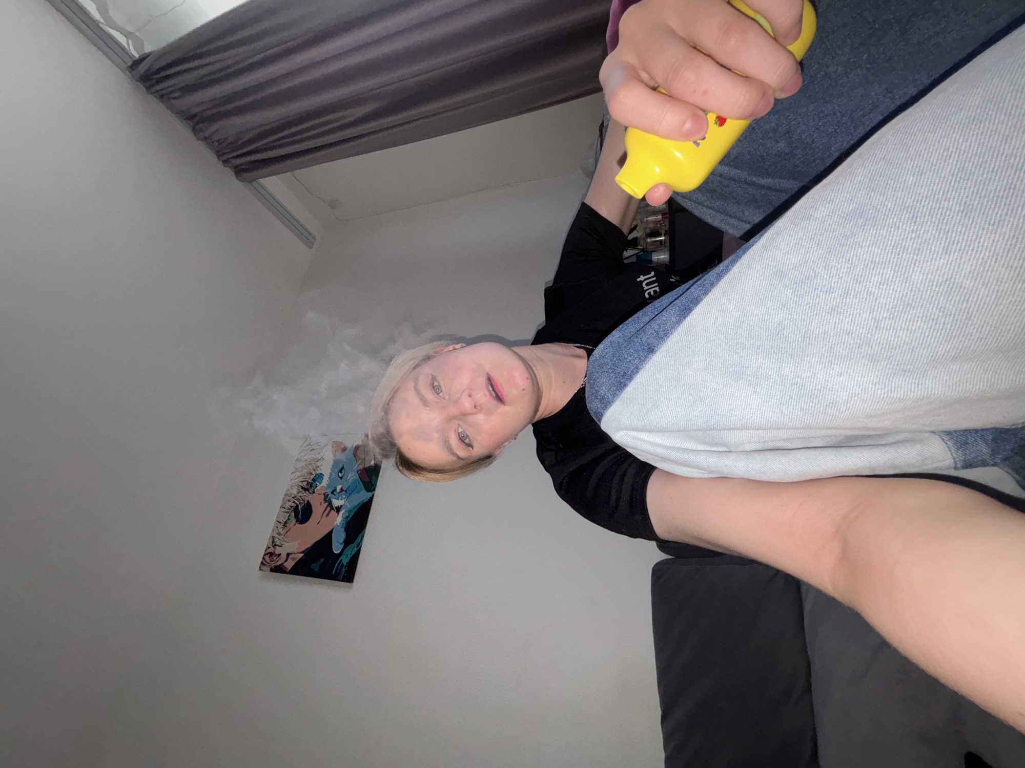 An indoor, low-angle shot captures a blonde individual with short hair, looking down directly at the camera. The person is positioned in a room with light-colored walls and a dark gray curtain or blind visible at the top. They are wearing a black top, partially revealing the word "ant" (reversed), and light blue denim jeans over a darker blue fabric, possibly another pair of jeans or a blanket. Their right hand holds a bright yellow, presumably plastic, bottle or spray device with small red and blue character-like designs.
A cloud of white vapor or mist emanates from the area around the person's face, suggesting they are either vaping or using a spray product. The person's expression is neutral to slightly curious. On the wall to the left, a colorful, stylized picture or painting of a blue creature, possibly a cat or fox, with prominent eyes, is visible, partially obscured by the mist. A dark gray piece of furniture, likely a sofa or bed, supports the person's lower body, with their leg in black socks resting on it. The lighting is internal, offering no specific cues about the time of day or outdoor weather. The scene depicts an individual engaged in an activity involving a handheld device and mist production within a private indoor setting in an unknown location in Russia.