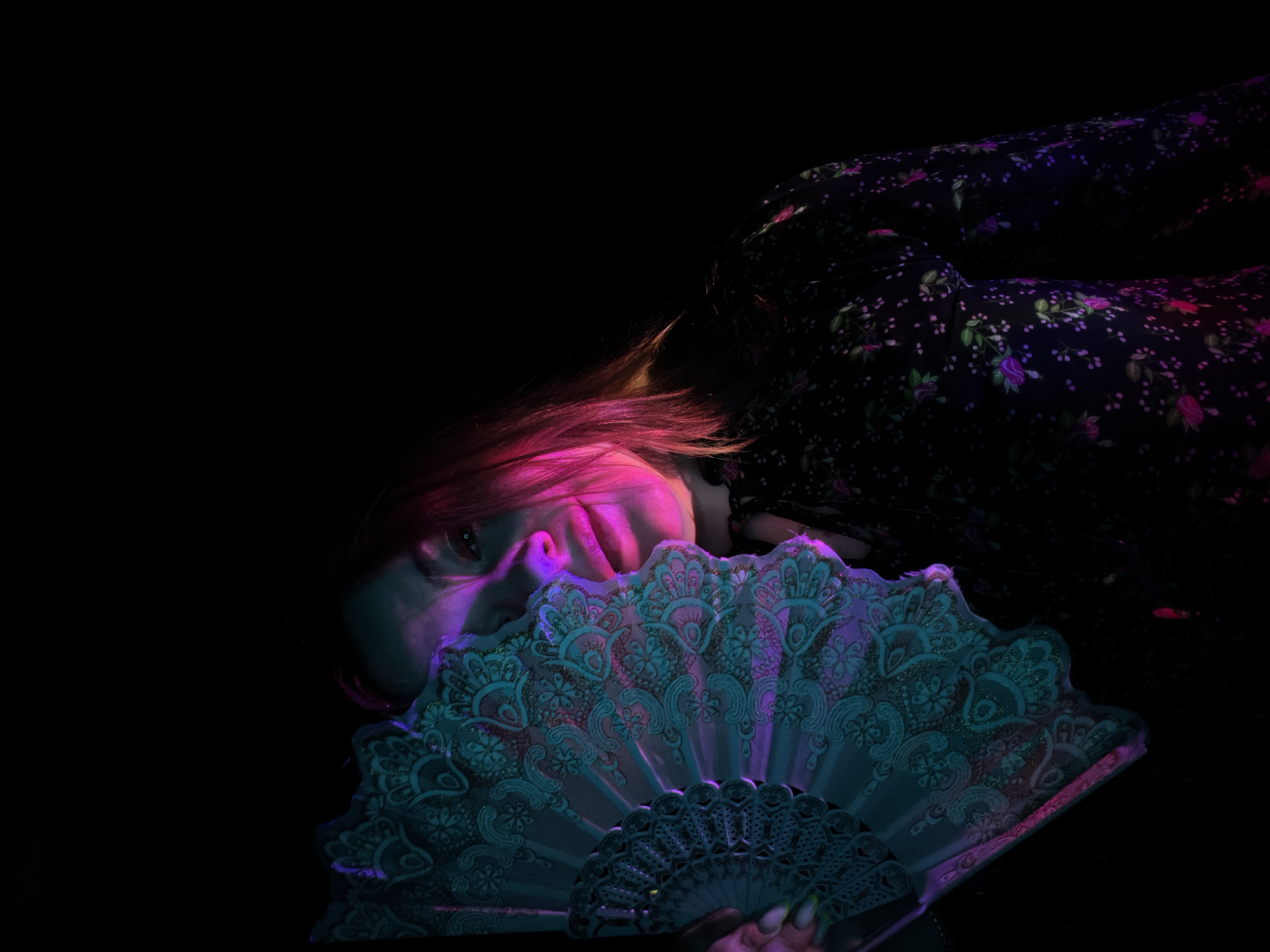 A woman lies in a dark area. She is partially obscured by an open fan with an ornate pattern. The woman's hair is highlighted with a pink hue. She is wearing a dark floral-patterned dress. The scene is illuminated by blue and purple light. The location is unknown, not specified as Chaikovsky, Russia.