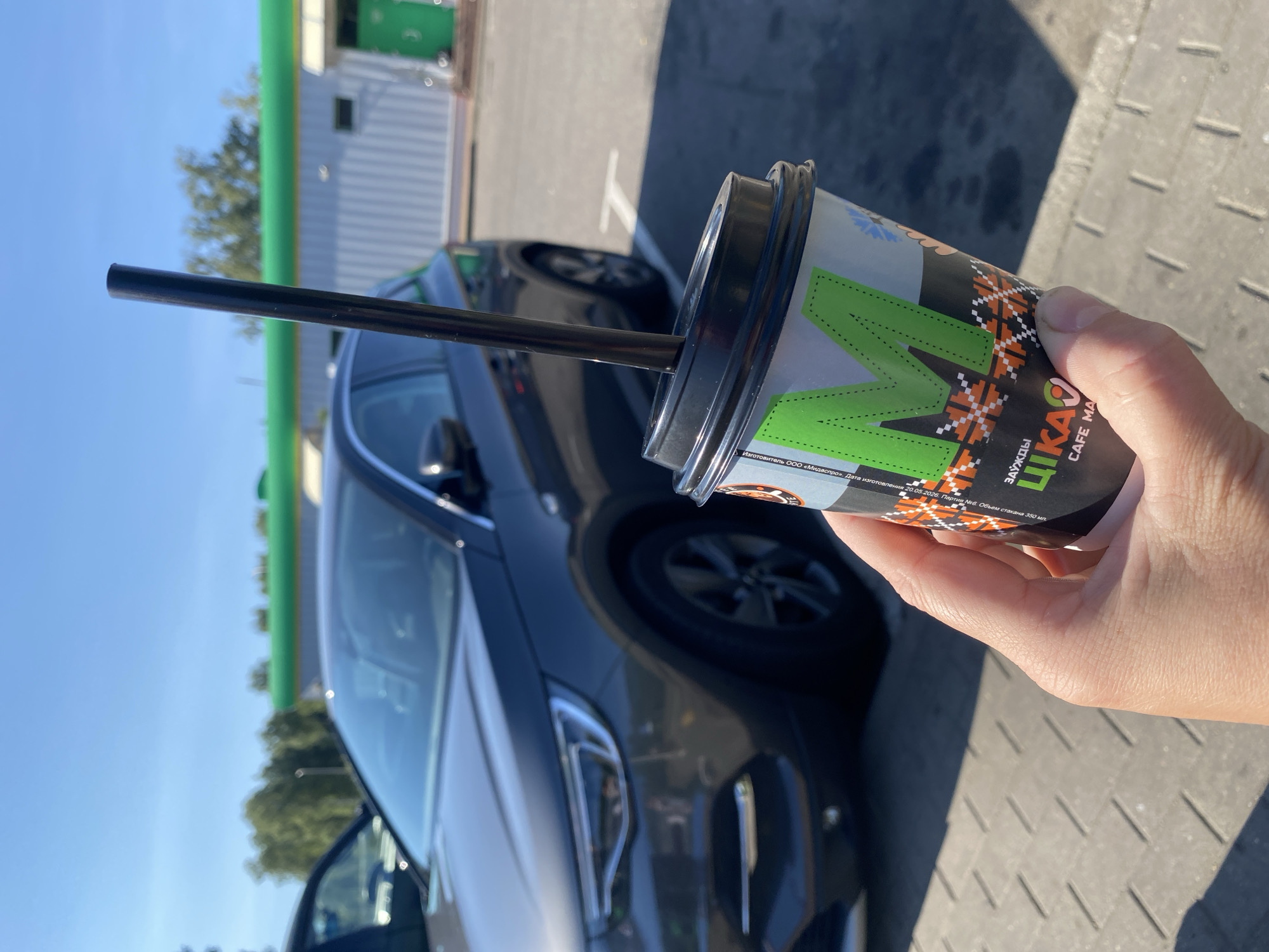 A person's hand holds a disposable coffee cup with a black lid and straw in the foreground, at what appears to be an outdoor gas station or rest stop. The cup has a colorful, patterned design with the letter "M" prominently displayed in green. In the background, a dark gray car is parked, and beyond that, a white building with a green roof is visible. The ground is paved with gray interlocking bricks, and there is a shadow cast by the cup and straw. The sky is clear and blue, suggesting a sunny day. The location is identified as Неманіцкі сельскі Савет, Belarus.