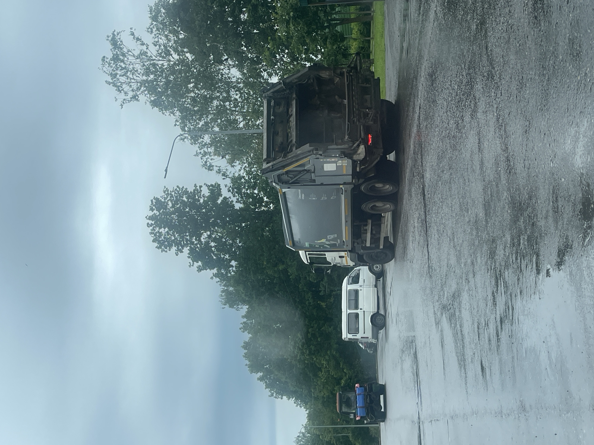 In Shakhovskaya, Russia, on a wet, overcast day, a garbage truck is parked on a paved surface. The truck, a large vehicle with a dark gray compactor body and a white cab, is positioned with its rear facing the viewer, revealing its open loading mechanism. To the left of the garbage truck, a white minivan is parked. Further to the left, a blue tractor with a red cab is also stationary. The ground is reflective due to recent rainfall, with visible puddles and streaks of water. Lush green trees form a dense backdrop behind the vehicles. The sky is a uniform gray, indicating cloudy weather. There are no people visible in the image.