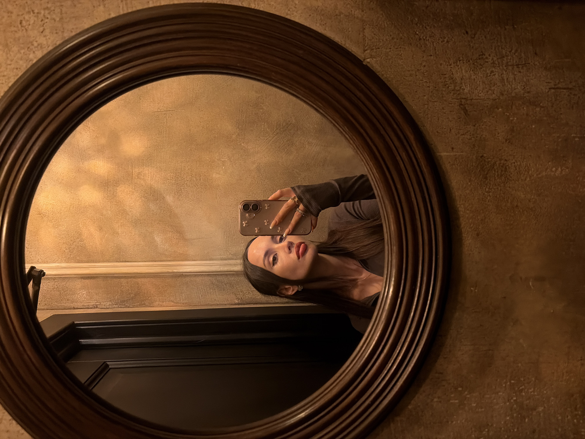 A woman is reflected in a round, dark-wood framed mirror, presumably taking a selfie with her smartphone, which has a patterned case. The reflection includes a glimpse of a dark cabinet or door below her. The background appears to be a textured, beige or tan wall. Based on the style of the mirror and the interior decor, the image may be located in Saint Petersburg, Russia.