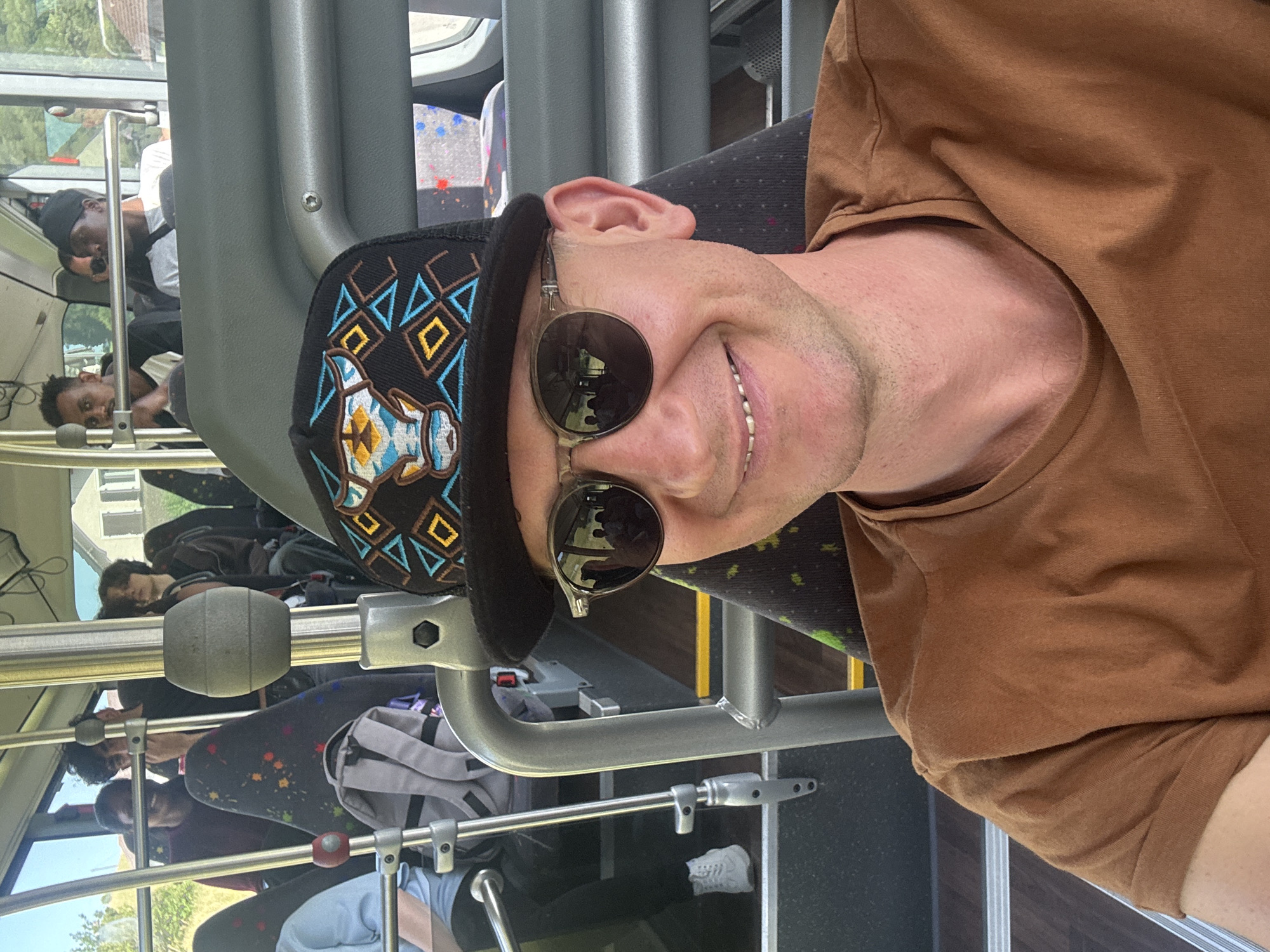 A close-up, selfie-style photo is taken from inside a bus in Saicourt, Switzerland, by a man with fair skin and a light beard wearing sunglasses and a brown t-shirt. He is also wearing a black baseball cap with a bull design on it. The man is smiling and looking at the camera. In the background, other passengers are visible on the bus, as well as the interior of the bus with seats and handrails. The windows of the bus show a blurred view of the outside, with trees and buildings visible. The lighting suggests it is daytime.