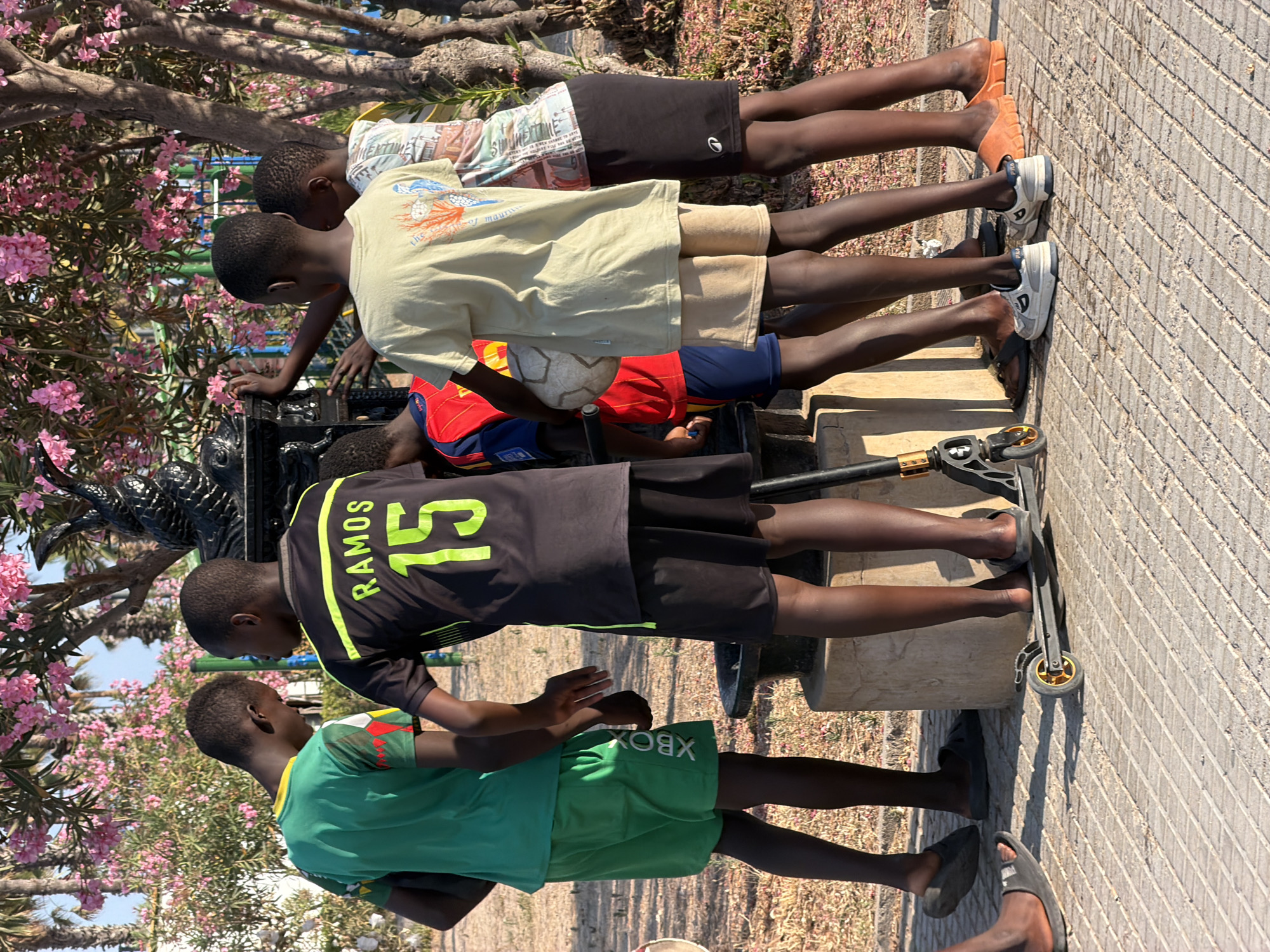 In Valencia, Spain, a group of six young boys are gathered near a fountain under a flowering tree. The boys, all appearing to be of African descent, are dressed in casual sportswear. Two of the boys are wearing soccer jerseys: one in green with red and yellow accents, and another in black with "RAMOS 15" printed in neon yellow on the back. Another boy is wearing a red and blue soccer jersey and holding a soccer ball. Two boys are wearing shorts, one pair green with "XBOX" written vertically, and another pair tan. One boy is wearing a patterned t-shirt and dark shorts. Two boys are standing on a scooter. The ground is paved with rectangular tiles. The environment is sunny with pink flowers visible on the trees.