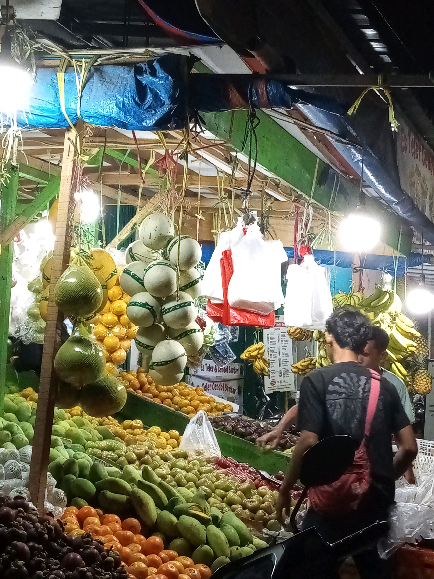 An outdoor market stall in Bekasi, Indonesia, is filled with a variety of fresh fruits. Melons, grapefruits, oranges, mangos, and bananas are prominently displayed, some hanging from the ceiling and others piled on shelves. The stall is covered with tarps and illuminated by bright lights, creating a bustling nighttime atmosphere. Two men are visible in the foreground, seemingly customers or vendors. One man, wearing a black t-shirt and a red sling bag, is looking away from the camera. The other man, wearing a green shirt, is partially visible behind him. The market appears to be busy, with abundant produce and a lively ambiance.