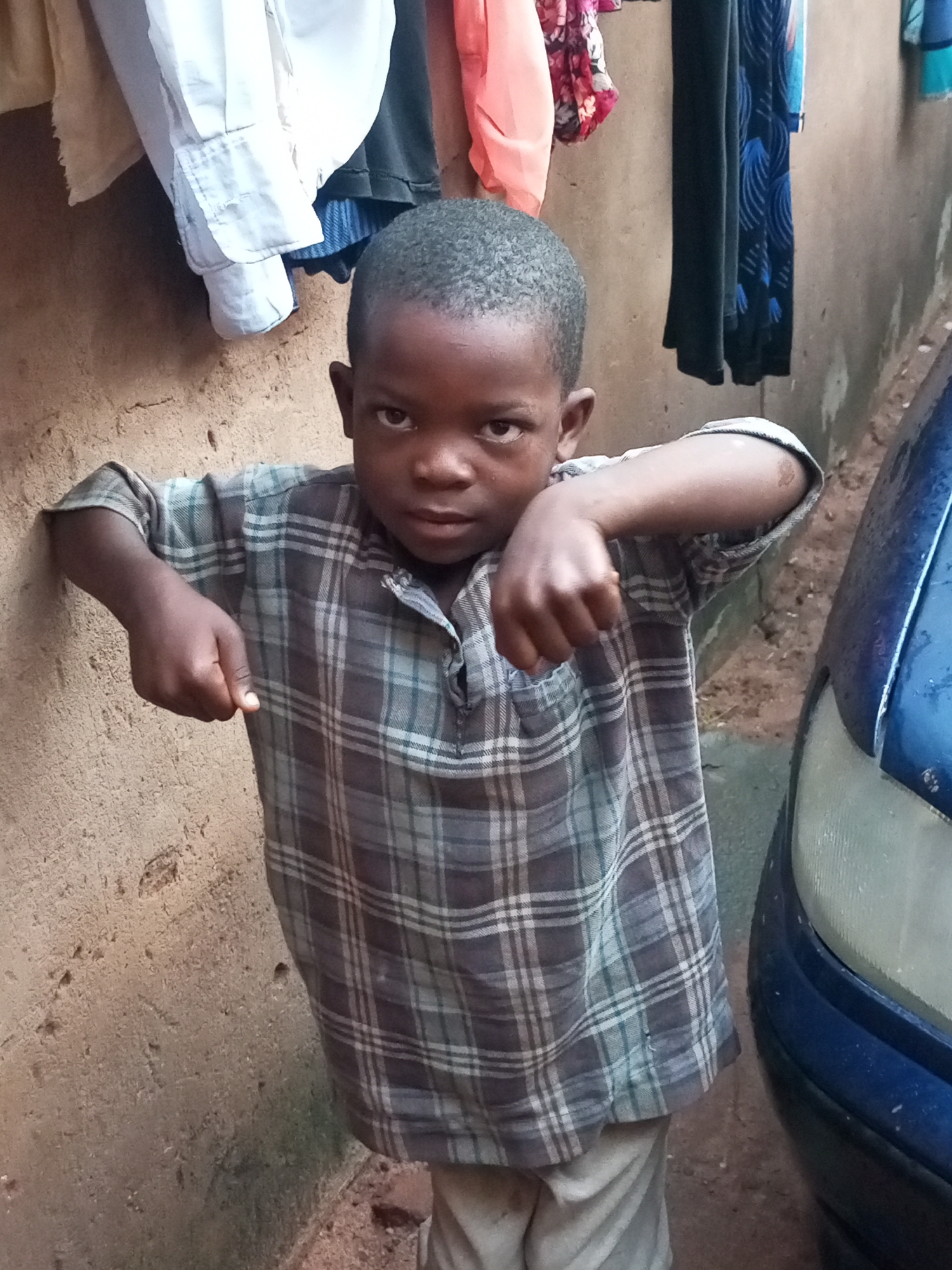 A young boy stands with his left arm bent and hand clenched, leaning against a rough, sandy-colored wall. He is wearing a plaid shirt and light-colored pants. The background shows clothing hanging on a line, suggesting a residential or communal outdoor area. To the boy's right, the edge of a blue car is visible, including its headlight. The scene appears to be in an outdoor setting in Tempure, Nigeria.