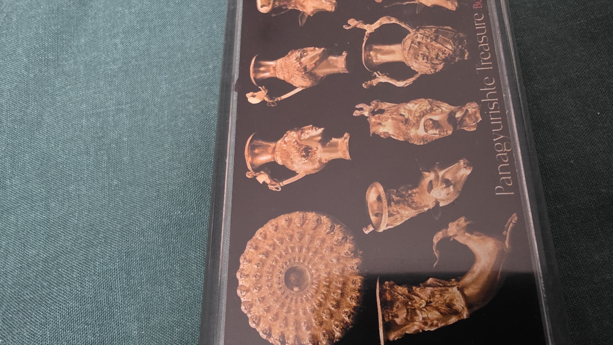 The image displays a close-up view of intricate gold artifacts, likely ancient, set against a dark background. The right side of the frame features these items, which include several ornate vessels such as pitchers or cups with animal head motifs, a large circular object resembling a patterned shield or plate, and a horn-shaped rhyton ending in an animal leg. The artifacts exhibit a rich golden color, suggesting they are made of gold or are gold-plated.
Vertically aligned along the right edge of the display, the text "Panagyurishte Treasure Bu" is visible. The "Bu" likely refers to Bulgaria, tying into the well-known Panagyurishte Treasure, a famous set of Thracian artifacts.
The display containing the artifacts appears to be resting on a textured, teal-green fabric surface, which occupies the left side and a small part of the bottom right of the photograph. No people are visible, and no specific event or activity is depicted beyond the presentation of these historical objects. The image provides a detailed look at the artistry and craftsmanship of the treasure, set within a general context of Blagoevgrad, Bulgaria, where such cultural heritage is often exhibited.