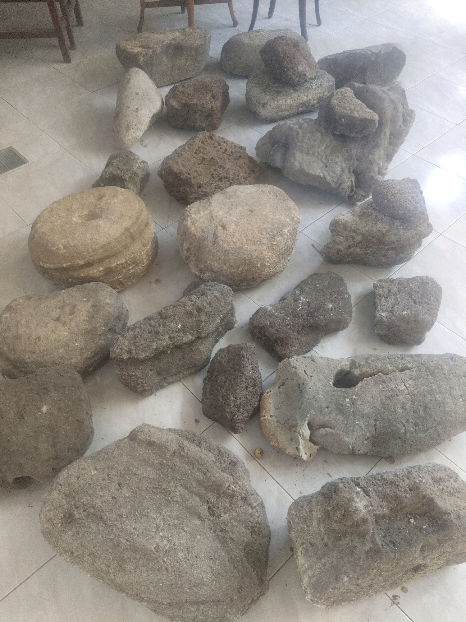 The image displays a collection of various-sized and shaped rocks scattered across a tiled floor. The rocks are predominantly gray and brown, with some exhibiting rough, porous textures while others are smoother and more rounded. One rock in the upper left appears to be a millstone with a circular indentation. The scene is well-lit, suggesting it is an indoor setting, possibly a workshop or storage area, with glimpses of wooden furniture legs in the upper portion of the frame. There are no people or animals visible in the image. The location is identified as Cisaga, Indonesia.