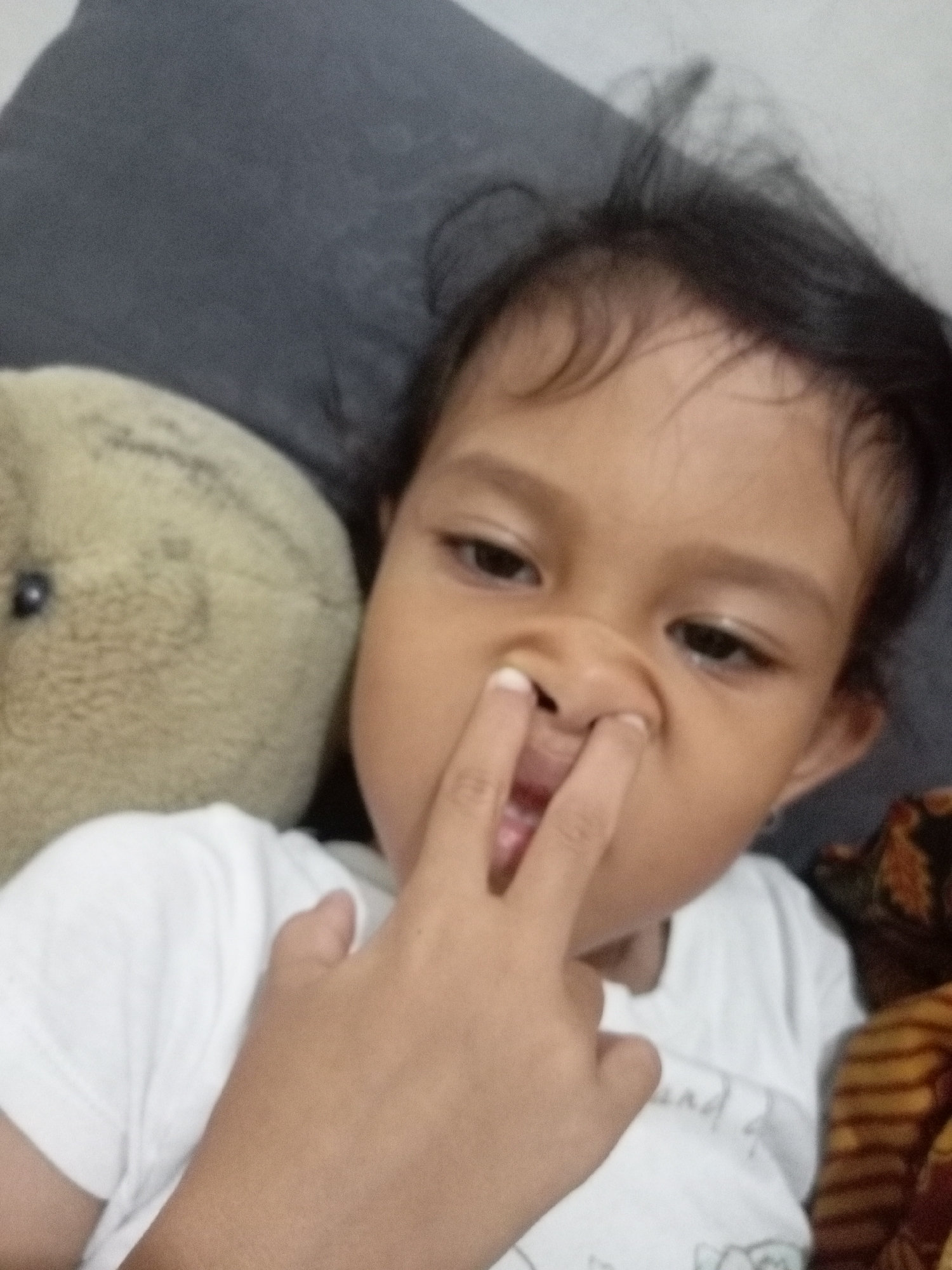 The image is a close-up, eye-level, indoor selfie of a young child, likely under the age of five, with dark hair. The child is lying down, with their head resting on a dark gray pillow. To the left of the child is a light brown, fuzzy stuffed animal, possibly a bear. The child is wearing a white t-shirt. The child's right hand is raised, with their index and middle fingers inserted into their right nostril. Their mouth is slightly open, revealing their tongue. The background consists of the pillow, the stuffed animal, and a blurry patterned fabric to the right. The lighting appears to be from an overhead source. The image does not provide any information about the location in Batam, Indonesia, or any specific environmental details that would suggest this.