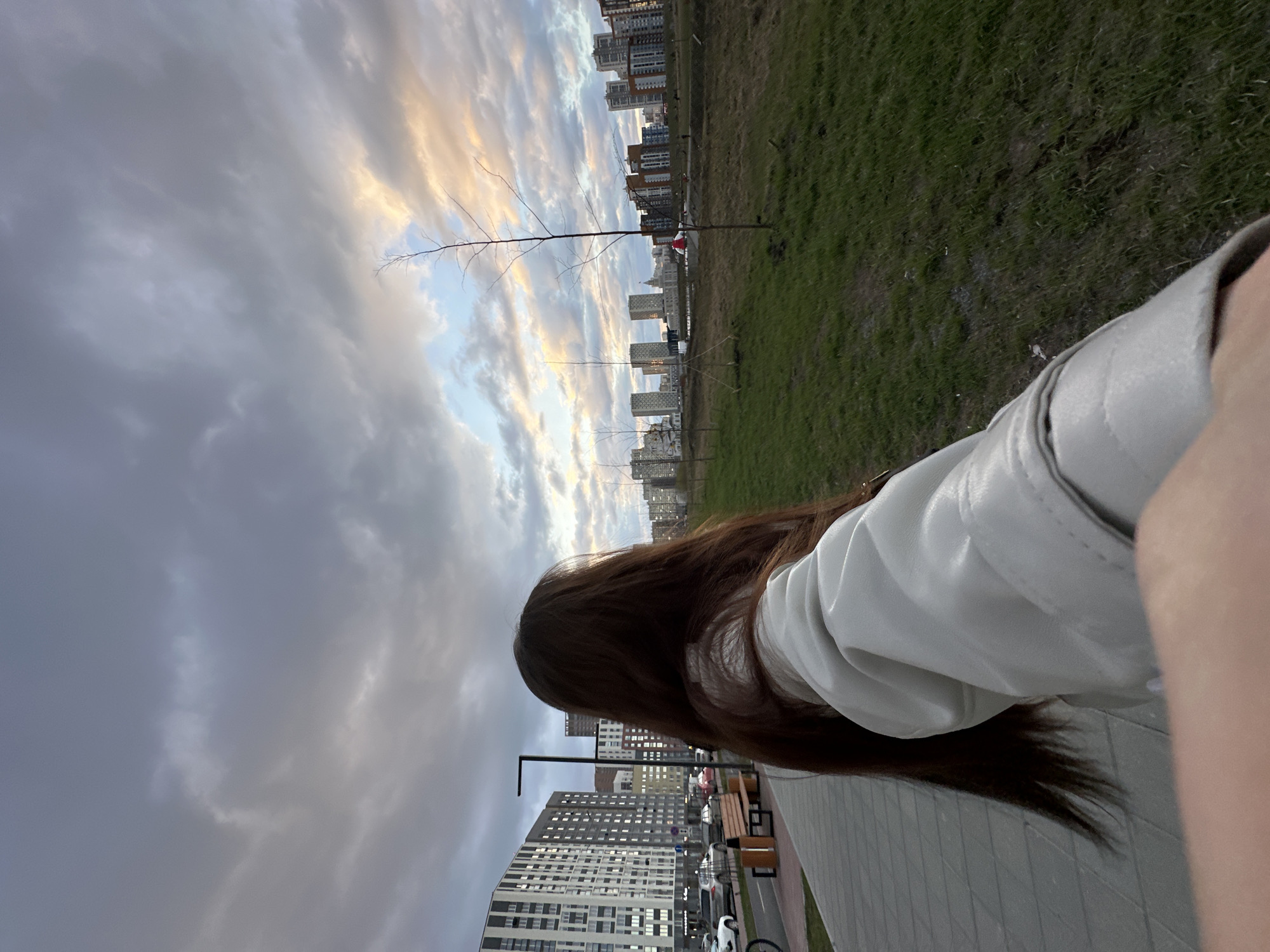 A person stands facing away from the camera, with brown hair and wearing a white jacket. The location appears to be a park or walkway within Yekaterinburg, Russia, given the presence of modern apartment buildings, benches, streetlights, and what seems to be a paved walkway with a bicycle. Buildings and green spaces are in the background beneath a clouded sky, possibly near sunset or sunrise. The photo seems taken from the first-person perspective.