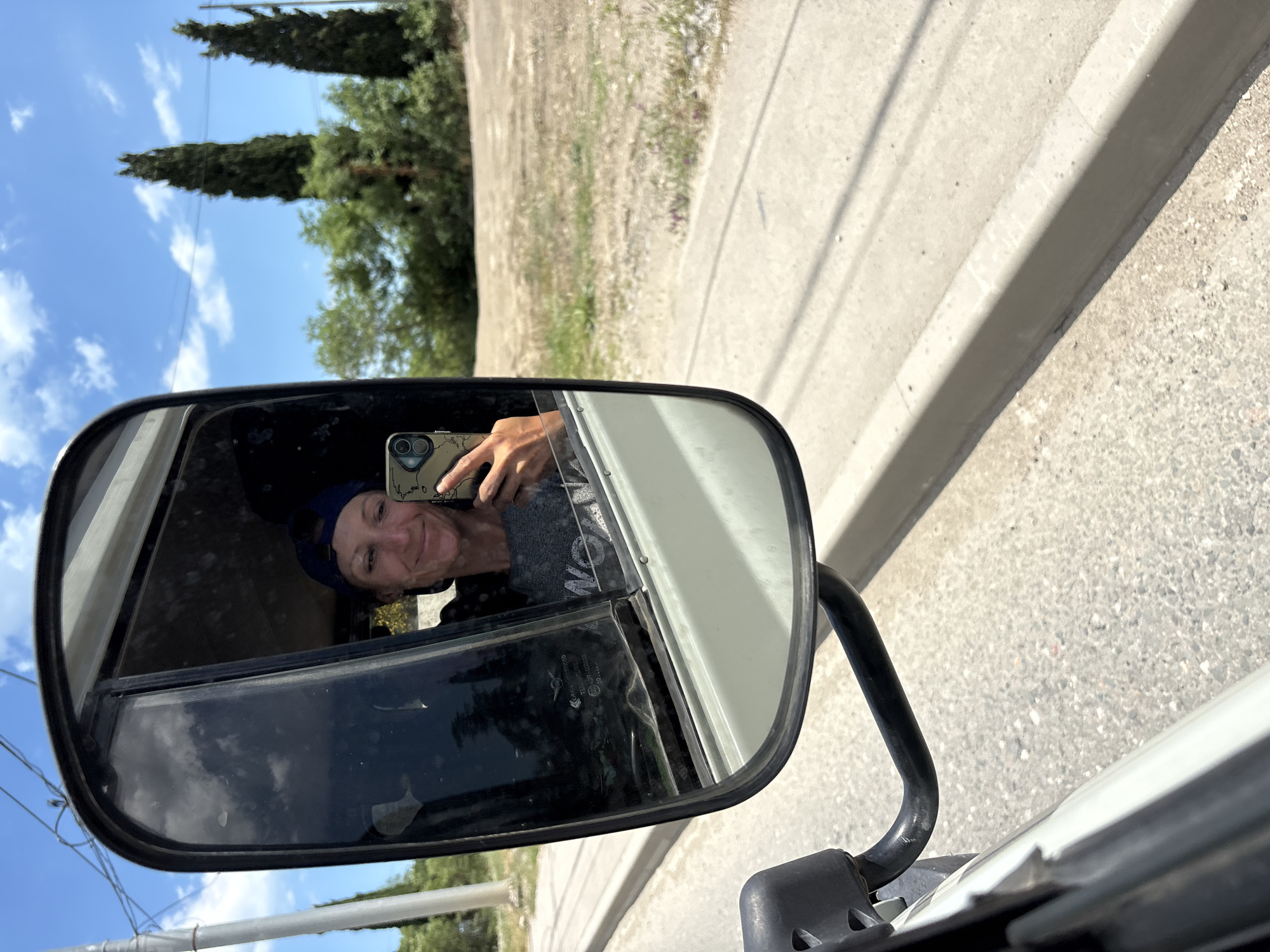 A woman wearing a blue baseball cap and a grey t-shirt is reflected in a truck's side-view mirror. She is holding a mobile phone in her right hand, taking a selfie. The background of the reflection shows trees and a blue sky with clouds, suggesting an outdoor environment. The truck appears to be in motion, as indicated by the blurry road and curb visible outside the mirror. The scene is set in Kiparisnoe, Ukraine.