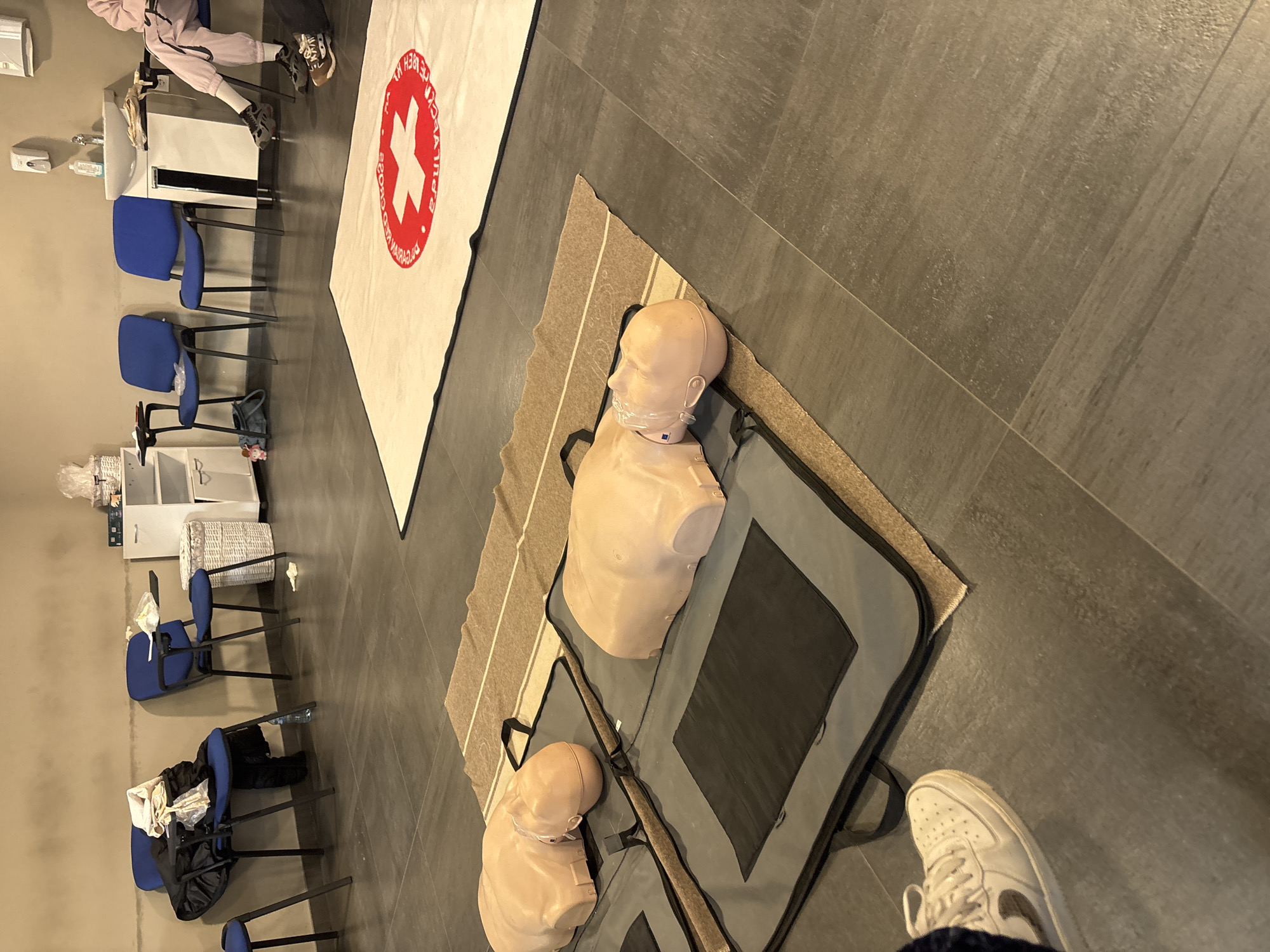 The image shows an indoor scene, likely a training room in Plovdiv, Bulgaria. The room has a gray tiled floor. There are rows of blue chairs along one wall. On the floor, there is a white banner with a red cross. Next to it are two CPR mannequins on a tan-colored mat. The mannequins are packed in grey carrying cases. A person wearing white sneakers is partially visible in the foreground. The scene suggests a first aid or CPR training session.