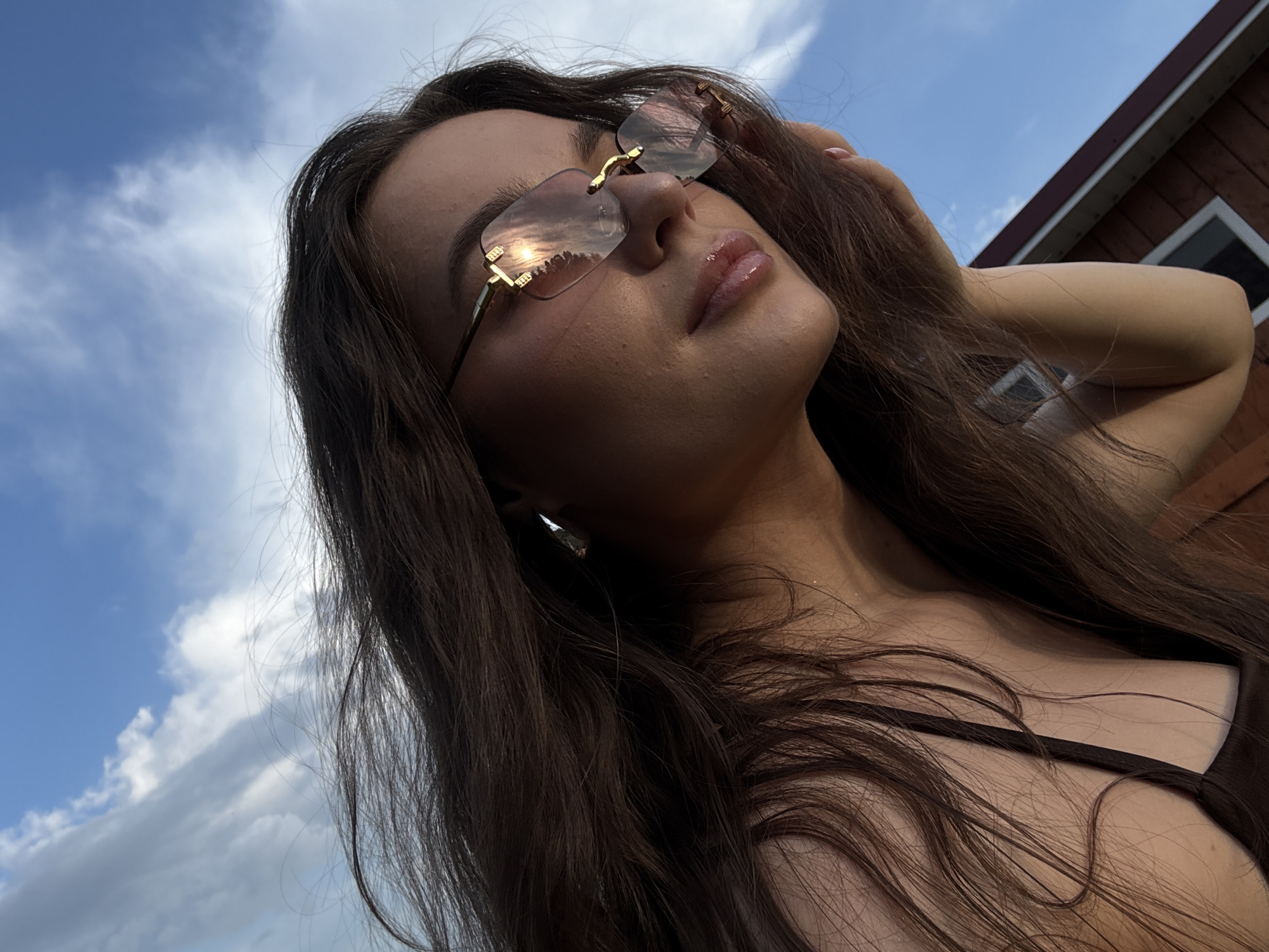 A young woman with long, dark brown hair, wearing oversized, rimless sunglasses with gold accents, is captured in a close-up, side-profile shot. Her eyes are closed, and her hand is gently resting on her cheek. The background is a clear blue sky with scattered white clouds. The reflection on her sunglasses shows trees and the sun. A small portion of a wooden building is visible in the lower right corner of the frame. The lighting suggests it is either early morning or late afternoon. The scene is likely outdoors, possibly in a rural or suburban setting, with no clear indication of the city of Rusalevka, Russia.