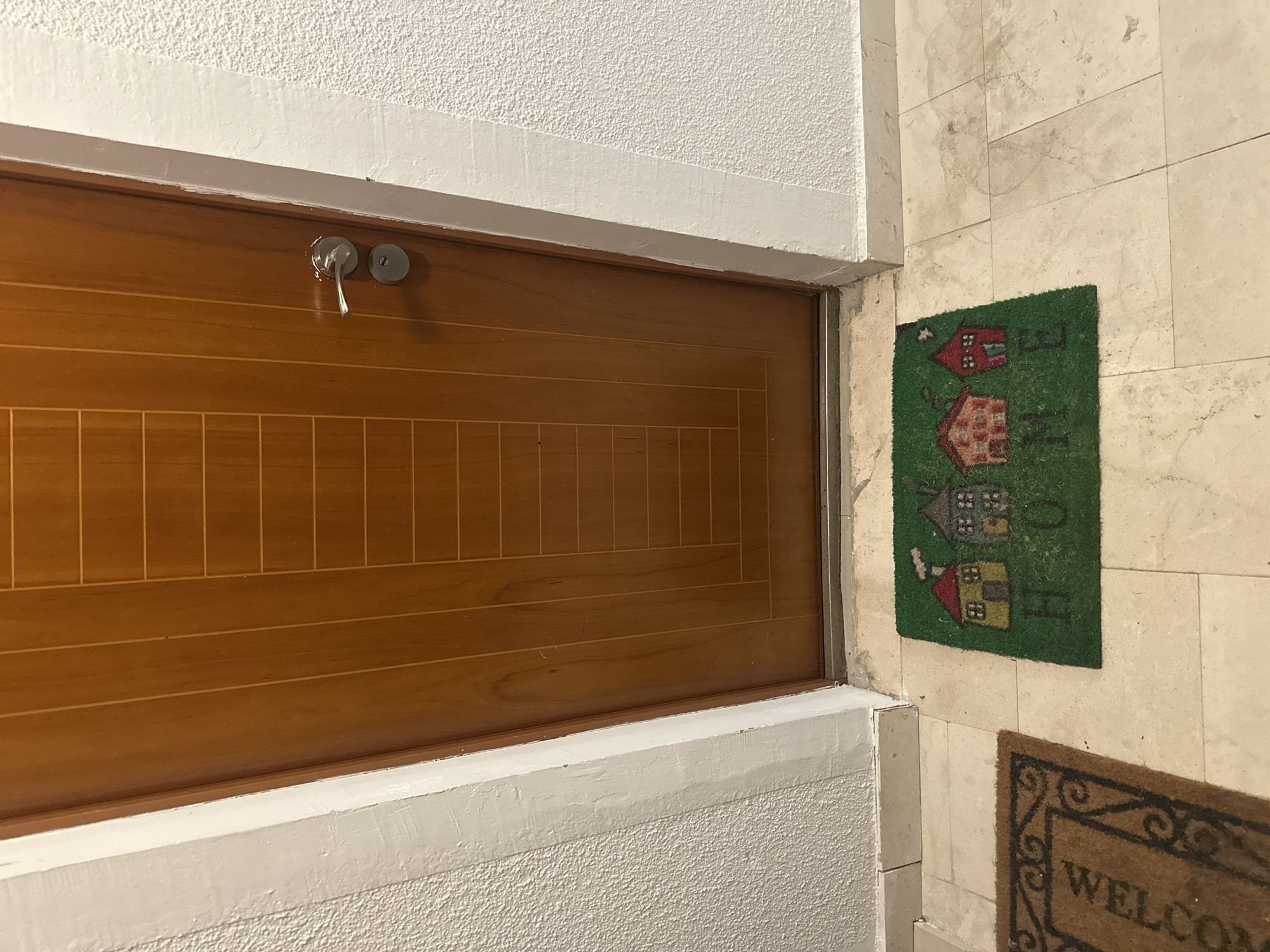 The image shows a portion of an interior entrance in Budva, Montenegro. A wooden door with a horizontal groove pattern is visible, with a key present in the lock. The door frame is painted white. Adjacent to the door, a green mat with depictions of houses rests on a light-colored tiled floor. A portion of another doormat with the word "WELCOME" is visible below. The wall is made of light-colored tiles. The ceiling is textured and painted white.