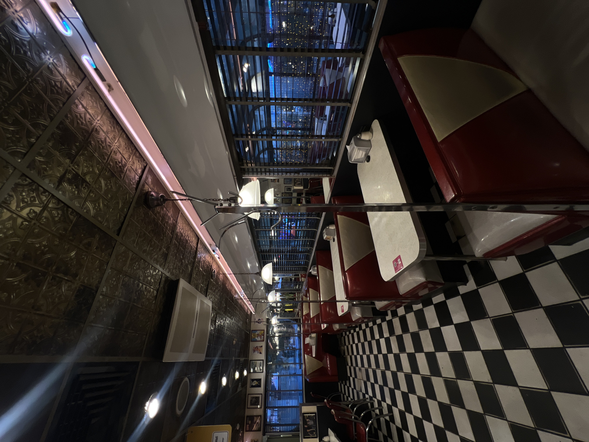 The image depicts the interior of a restaurant in Moscow, Russia. It is a typical diner setup with red and white booths lining the walls and a checkered black and white floor. Above the booths is a ceiling with a window that reflects the outside city lights. On the left wall, there is a decorative patterned wall with a pink strip light and framed photos near the back. The lighting fixtures are chrome with globe shades.