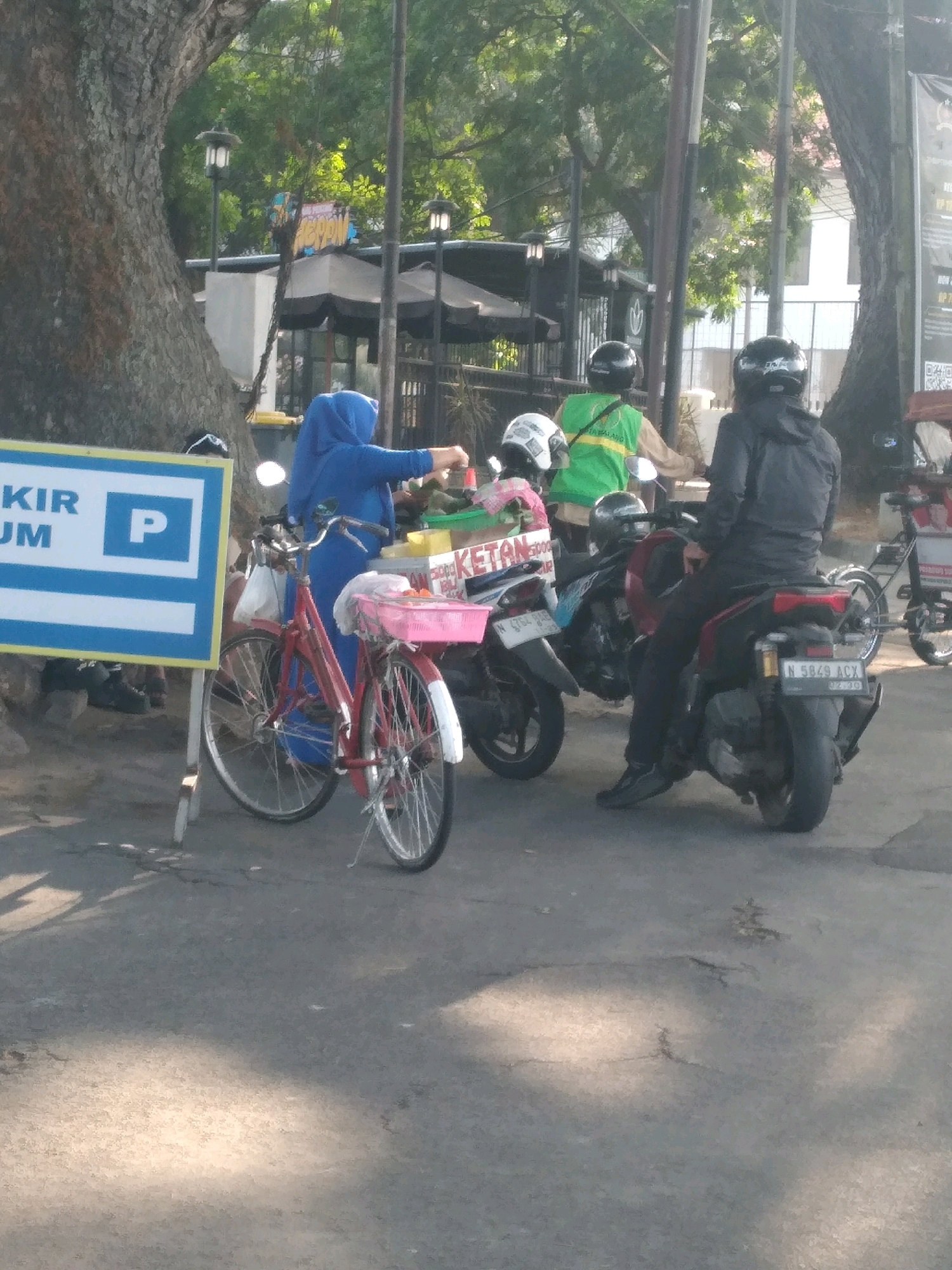 In Malang, Indonesia, on a street with a large tree, a woman in a blue hijab stands next to a red bicycle. She is reaching into a food cart labeled "KETAN," which is adorned with green and red boxes. The bicycle has a pink basket on the front, holding a white bag and some red items. Behind the woman and the bicycle, two men on motorcycles are visible. The man on the right wears a black jacket and helmet, his motorcycle has a license plate reading "N 5849 ACX". The man to his left wears a green vest and helmet. Another motorcycle is parked to the left of the food cart. A blue and white parking sign stands to the left of the bicycle. The ground is paved, with shadows of trees and light filtering through them.