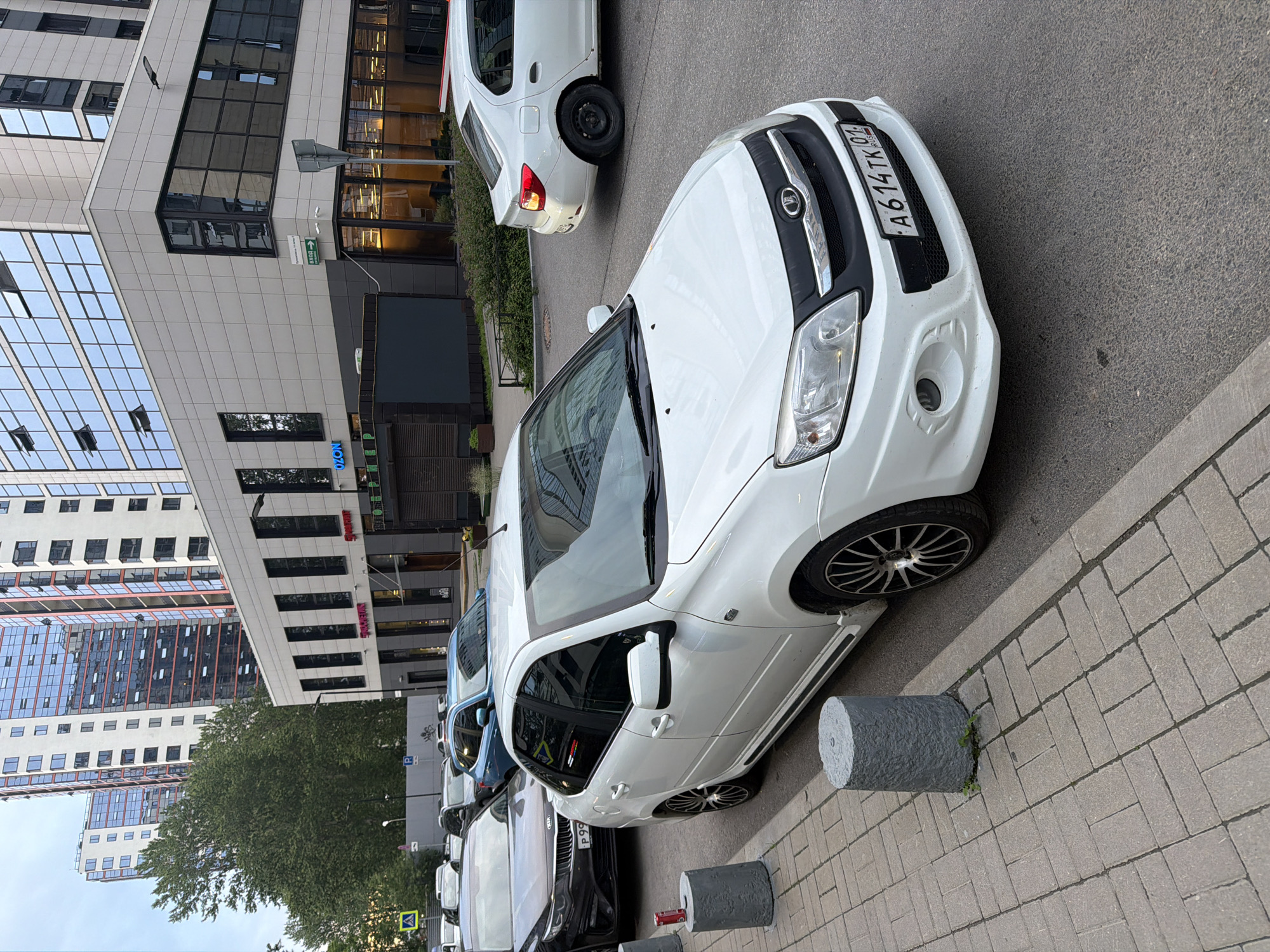 A white sedan is parked on a street in front of a modern building. The car is a Lada Granta, license plate A614TK 01. It is facing towards the viewer, with its front bumper and hood visible. To the left of the car, there is a sidewalk with three concrete bollards. In the background, there is a multi-story building with large windows and balconies, and some trees. The sky is overcast.