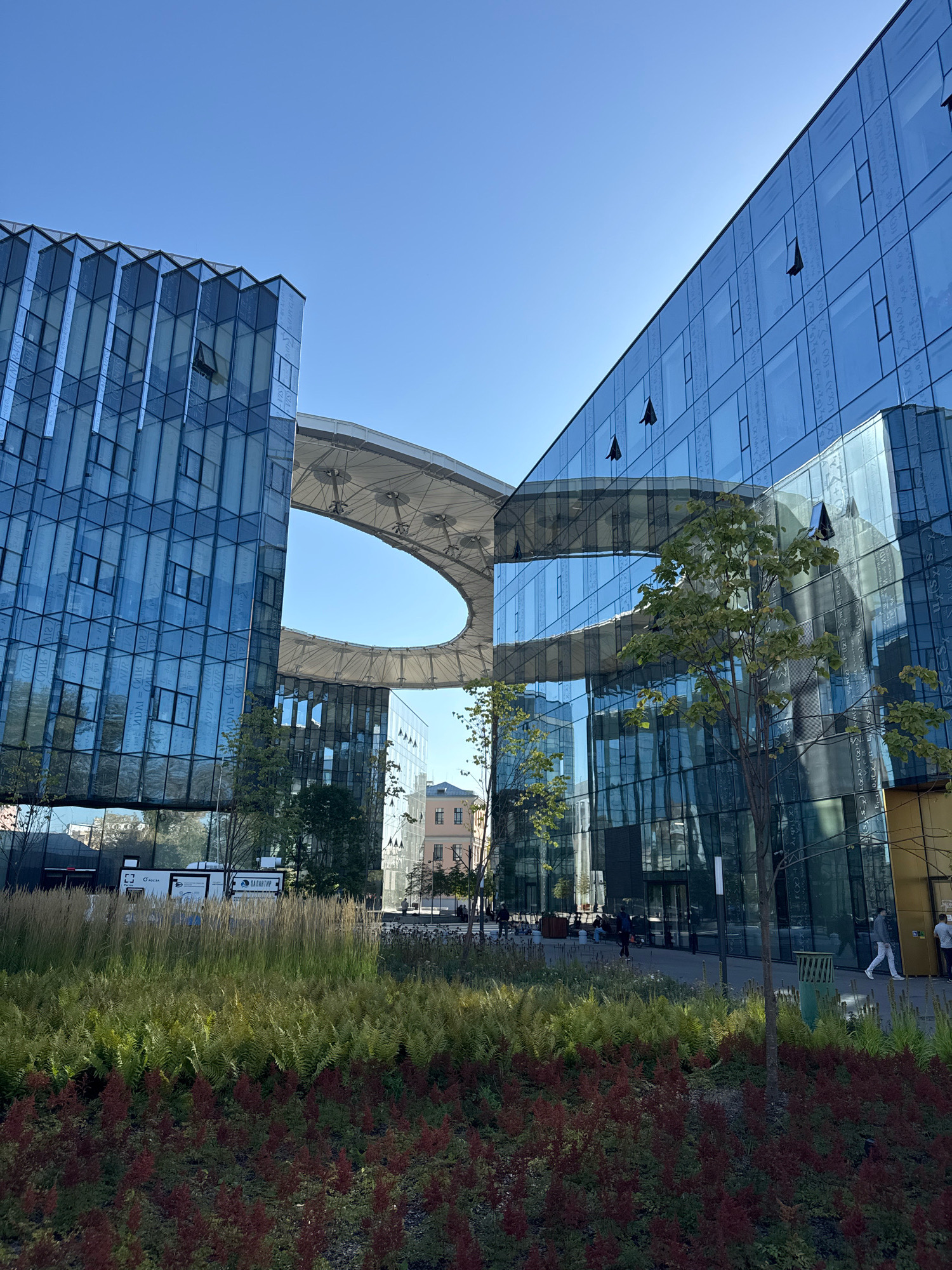 The image shows a modern architectural complex in Moscow, Russia, likely a business or innovation center, featuring sleek glass buildings. A prominent feature is a circular sky bridge connecting two of the buildings, creating an oval-shaped opening against the clear blue sky. The facades of the buildings are adorned with what appears to be mathematical formulas etched or printed on the glass, adding a unique aesthetic.
In the foreground, there is a well-maintained landscaped area with lush greenery, including tall grasses and ornamental plants with red foliage. Several people are visible in the mid-ground and background, walking or sitting around the plaza area, suggesting a public or semi-public space. One person is seen walking towards the right, while others are seated on benches. The overall impression is one of a contemporary urban environment that blends modern architecture with natural elements. The location is the Skolkovo Innovation Center.