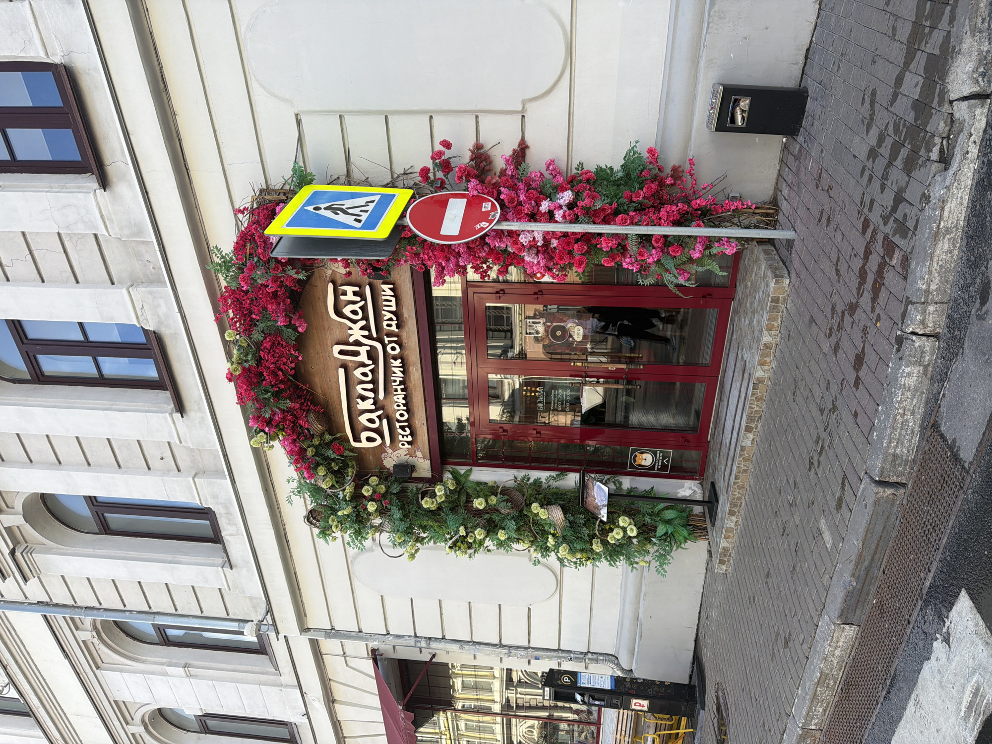 This is a vertical, eye-level outdoor photograph of a building facade decorated with abundant floral arrangements. The building is painted a light cream color and features several dark-framed windows. One prominent window is a dark red double door with glass panels, suggesting an entrance to a business.
The entrance is framed by a lush garland of artificial red and pink flowers intertwined with green foliage and branches. Above the door, a wooden sign with Cyrillic text is partially visible, reading "Баклажан" (Baclajan) and "ресторанчик от души" (a little restaurant from the soul).
To the left of the entrance, two road signs are mounted on a metal pole: a blue and yellow pedestrian crossing sign and a red and white "no entry" sign. A black trash receptacle is mounted on the wall to the right of the signs. The sidewalk in front of the building is paved with grey cobblestones, and a wet patch indicates recent rain or a damp surface. Further down the sidewalk, a metal grate suggests a drainage system.
The overall scene appears to be a street in an urban setting, likely a commercial area given the signage and business entrance. The time of day is difficult to determine precisely, but the lighting suggests it is daytime. The weather appears overcast or recently rained. No people are clearly visible. The vibrant floral decorations add a decorative and welcoming element to the building's exterior.