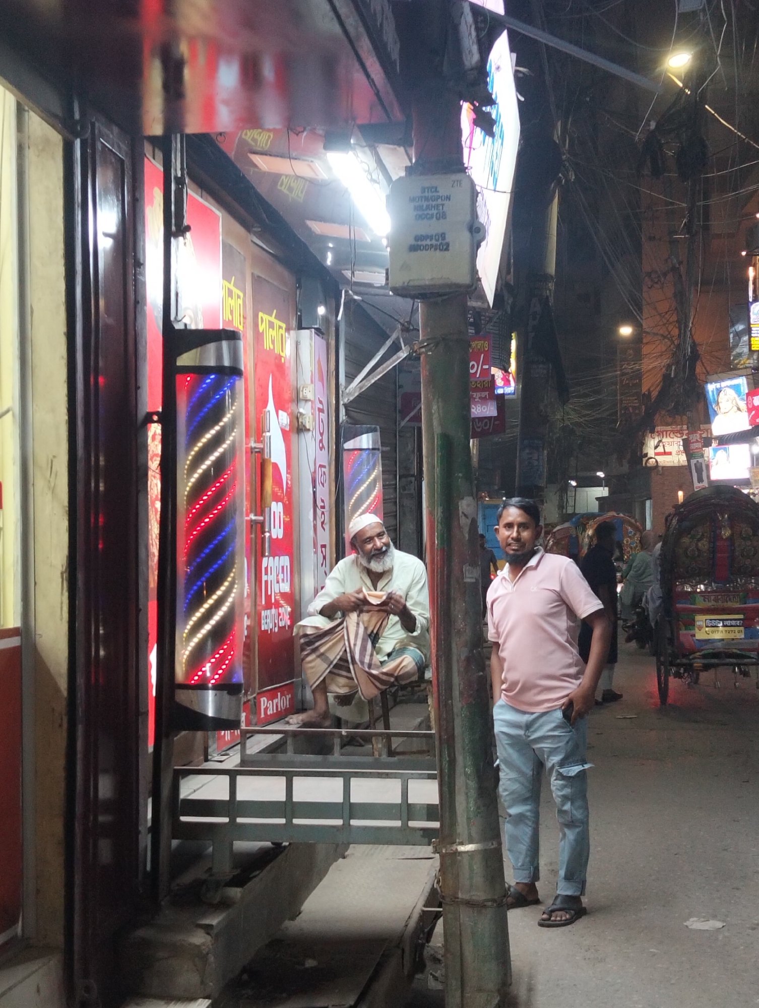 The image shows a street scene in Dhaka, Bangladesh, featuring a barber shop and a rickshaw. In the foreground, a man in a pink polo shirt and blue cargo pants stands casually with his hand in his pocket. Beside him, an older man sits on a small stool, wrapped in a traditional checkered cloth, appearing to eat something from his hand.
Behind them, the barber shop is visible with a classic spinning barber pole in front. Signage with Bengali script is also visible. Further down the street, a rickshaw is parked, and other pedestrians are present. The street is cluttered with overhead wires and various signs. The scene captures the bustling street life and the blend of traditional and modern elements.