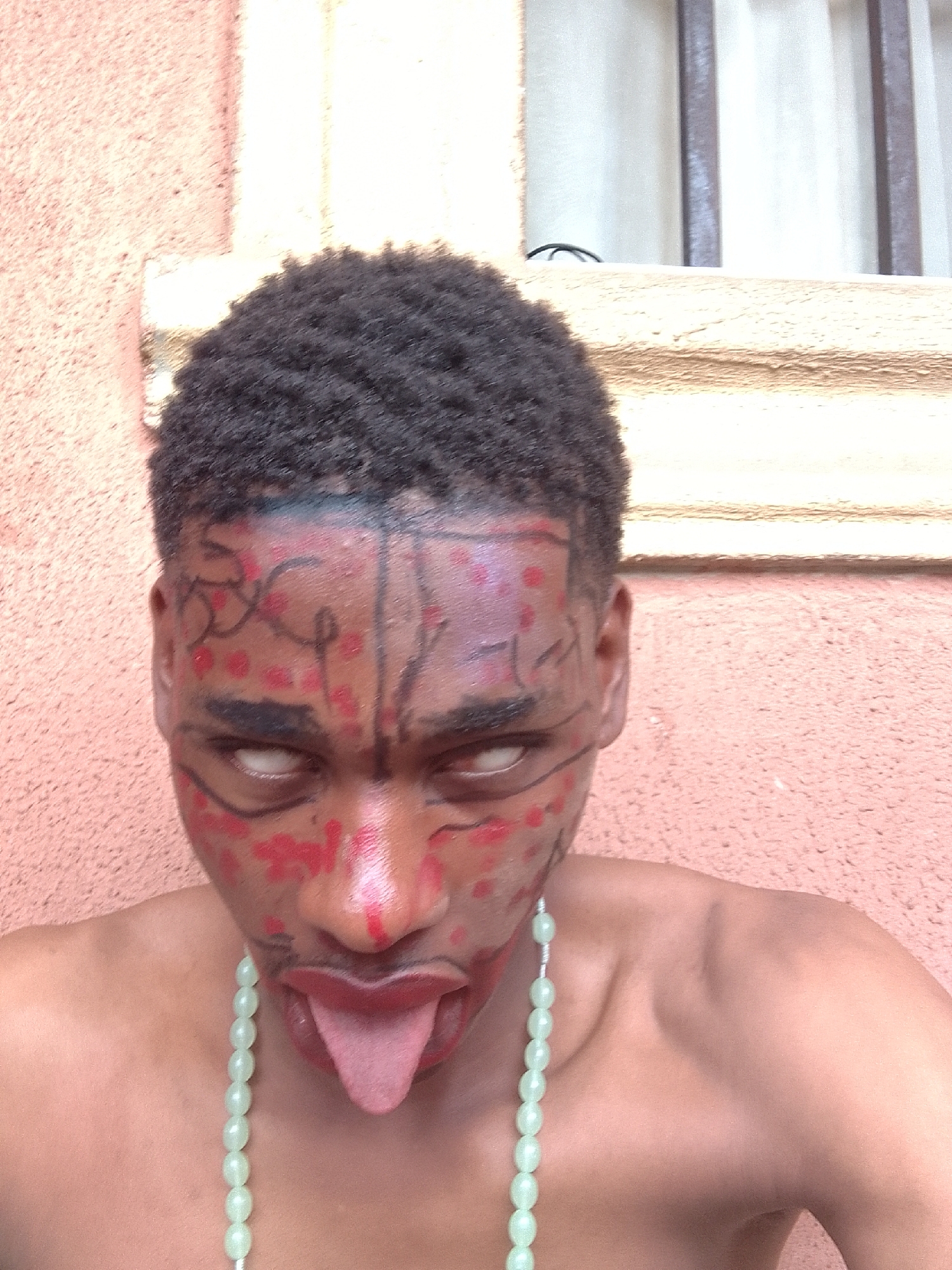 The image displays a young person, likely male, captured from the chest up, looking directly into the camera. He has short, dark, curly hair. His face is elaborately painted with bold black lines forming patterns across his forehead, cheeks, and around his eyes, combined with numerous red dots and smudges that resemble blood or ritualistic markings. His eyes are rolled back, revealing only the whites, creating an intense and somewhat eerie gaze. His tongue is extended, sticking out from his mouth, adding to the dramatic and expressive pose. Around his neck, he wears a light green beaded necklace.
The background consists of a textured wall, primarily in shades of peach and light brown, suggesting an outdoor or semi-outdoor setting next to a building. Above his right shoulder, a window with dark vertical bars and a white curtain behind it is visible. The lighting indicates it is daytime, with natural light illuminating the subject. There is no visible text in the image. The overall scene conveys a strong sense of artistic performance, character portrayal, or cultural expression.