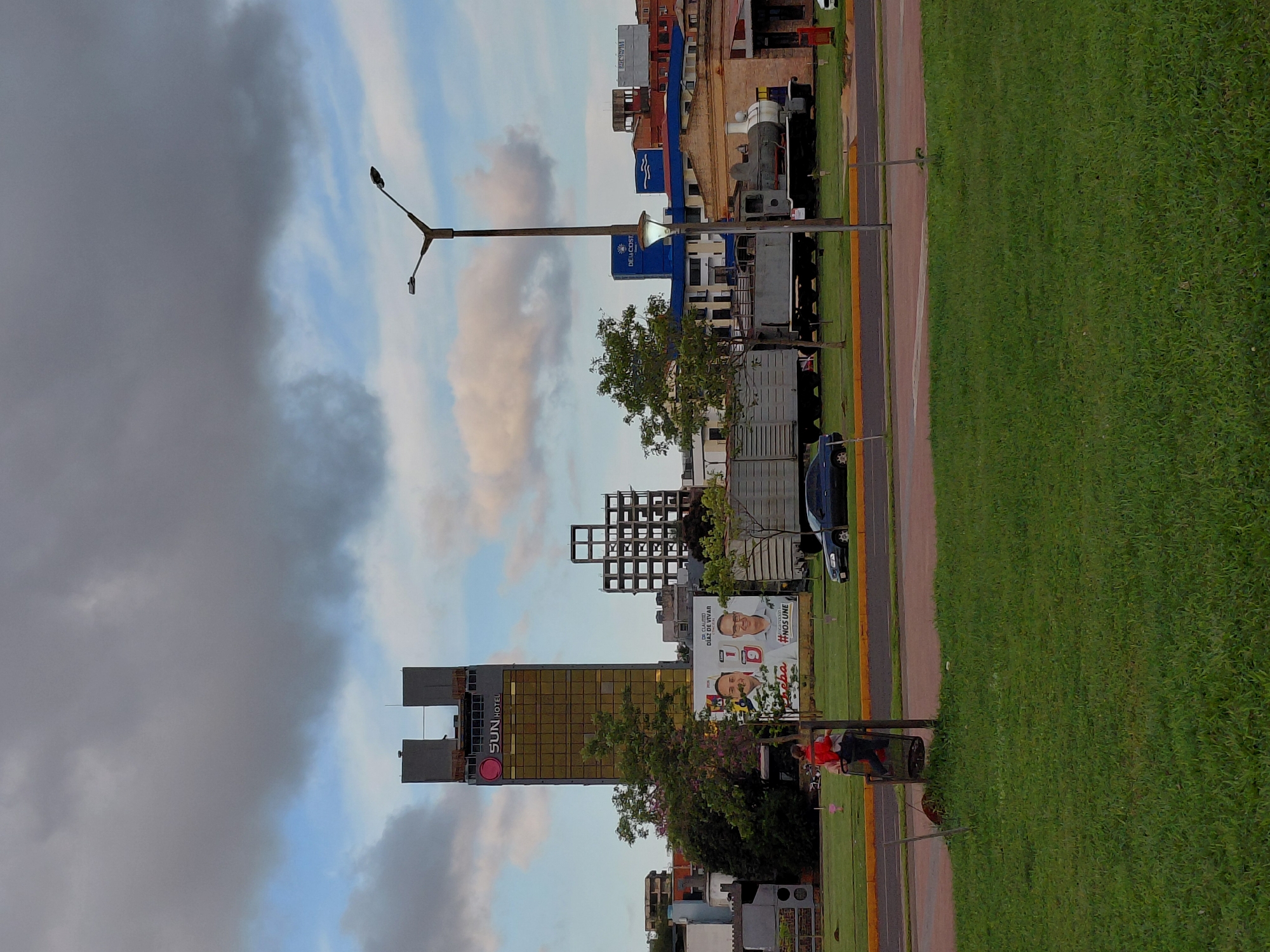In Encarnación, Paraguay, an urban scene unfolds under a cloudy sky. A prominent golden skyscraper, the "Sun Hotel," stands tall, its upper floors unfinished. To its right, a skeletal concrete structure, possibly another building under construction, pierces the sky. In the foreground, a well-maintained green lawn borders a paved area with a road. A woman in a red jacket pushes a cart, likely for waste disposal, near a trash receptacle. Across the road, a vintage steam locomotive and a freight car are displayed as static exhibits. Behind them, a row of buildings includes a blue structure with "Devacosta" branding and another with wave-like motifs. A street lamp with two lights stands on the right side of the image. A large political billboard featuring two men, one identified as "Dr. Claudio Diaz de Vivar," is prominently displayed near the hotel.