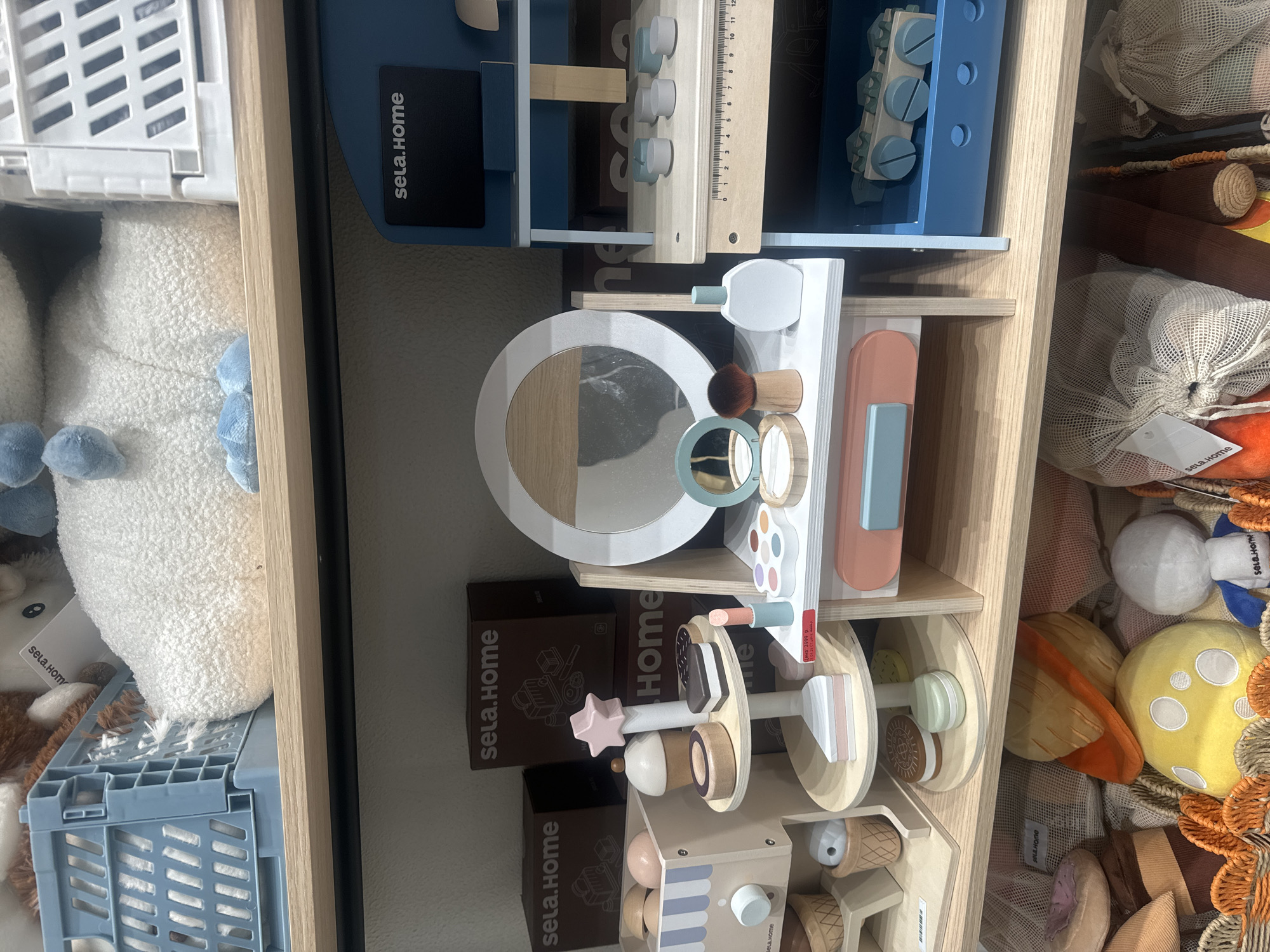 The image shows a shelving unit filled with toys and stuffed animals. The shelves are made of light-colored wood.
On the top shelf, there is a blue toy workbench with various tools. To the left of this is a white plastic basket containing a plush white animal toy with blue accents.
The middle shelf features a white toy vanity with a mirror, makeup, and a brush. Beneath the vanity are several brown boxes labeled "sela.Home".
The bottom shelf contains a toy ice cream stand with various ice cream cones and a pink star. To the right of this, there is a collection of stuffed toys, including a planet, a moon, and various other shapes, all contained within a mesh bag.
The image includes labels "sela.Home" on several of the boxes and toys, and a price tag of 3999 p.
Given the Russian text on the price tag, it is likely that the image was taken in the city of Krasnoyarsk, Russia.