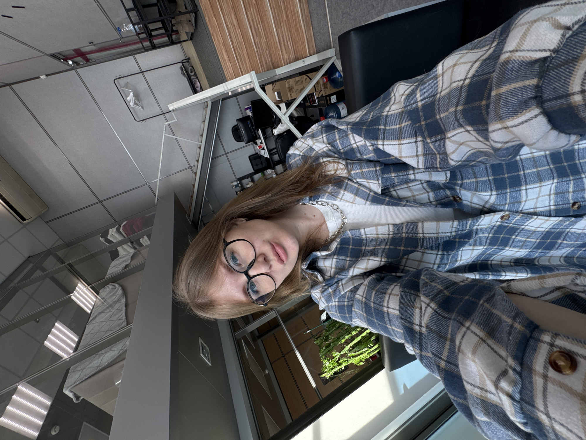 A young female with light brown hair and black-rimmed glasses is depicted taking a selfie indoors. She wears a white ribbed top, a silver-colored chain necklace, and a blue, white, and tan plaid button-up shirt. Her expression is neutral with slightly pursed lips.
The environment is a modern, multi-level commercial or office space located in Perm, Russia. The subject is seated, likely in a black chair, on the lower level. To her left, a large window or glass partition allows natural light to enter, and a tall, green, potted plant with multiple stems is visible. The ceiling is dark grey with recessed square light fixtures.
The upper level, accessible via a visible white staircase and framed by glass partitions, contains rolled-up items resembling mattresses or pads (light grey with a diamond pattern) and stacked textiles, including red, white, and black items. Fluorescent strip lighting is reflected in the upper glass panels.
The lower-level background, to the subject's right, reveals a workspace with grey paneled walls featuring diagonal lines and a mixed floor of grey carpeting and light wood-colored laminate. Visible objects include a black metal clothing rack holding at least two white hangers, shoes on the floor beneath the rack, and a black office desk with two black office chairs. The desk supports two computer monitors, white cups, and other small items. Further right, a black shelving unit holds multiple large plastic water bottles with blue caps, and several cardboard boxes are stacked, some bearing labels such as "ASUS" and "MEXICO." A white wall-mounted air conditioning unit is visible on the upper portion of the wall.