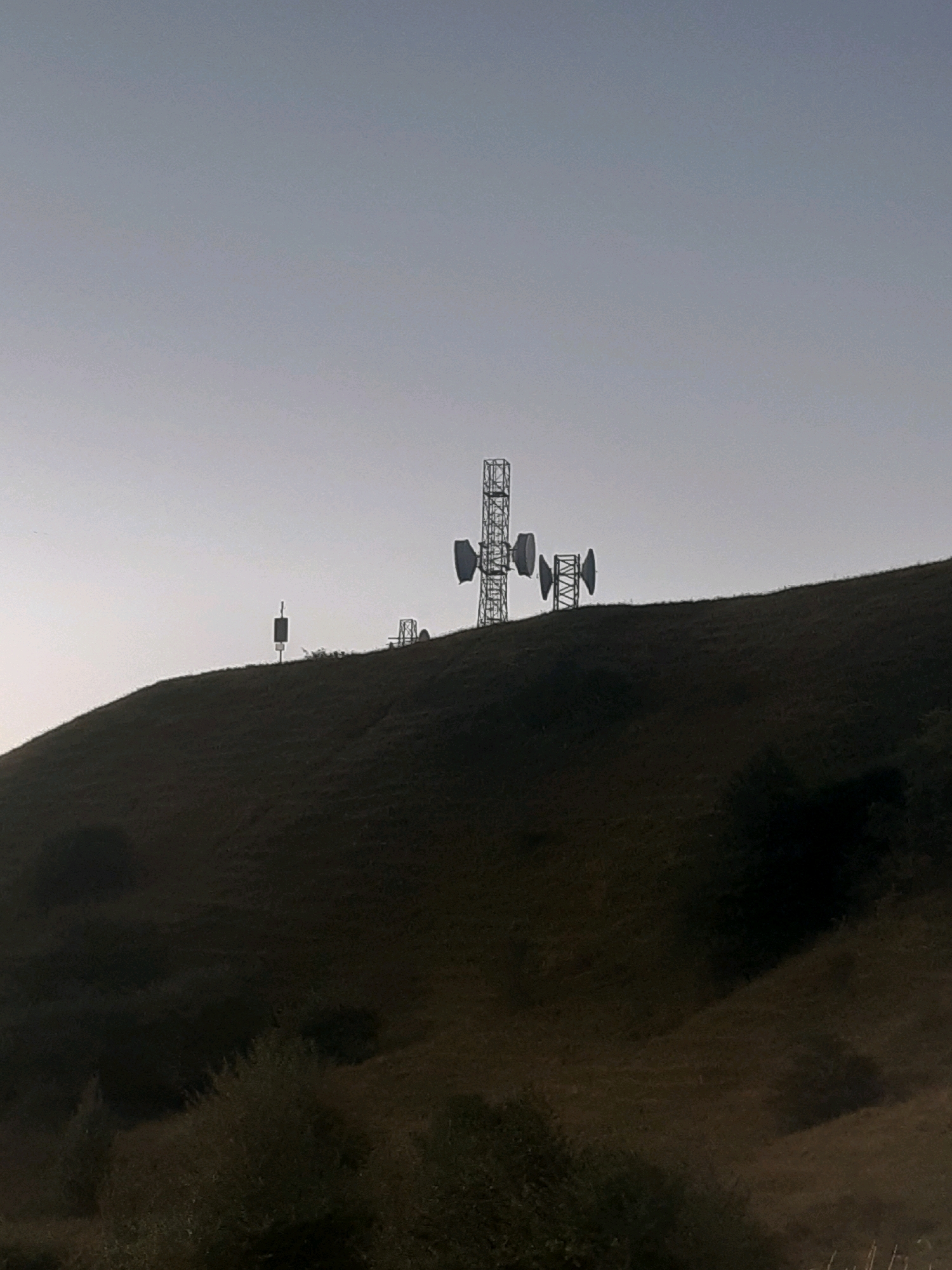 The image shows a silhouette of a communication tower on a hill. The sky is a pale blue and the hill is covered in dark vegetation, suggesting it might be dusk or dawn. There is no visible indication of the city of Verona, Georgia in the image. The communication tower is a tall metal structure with several antennas and dishes attached to it. There are also a few smaller structures and antennas on the hill. The overall scene is one of a rural or remote area.