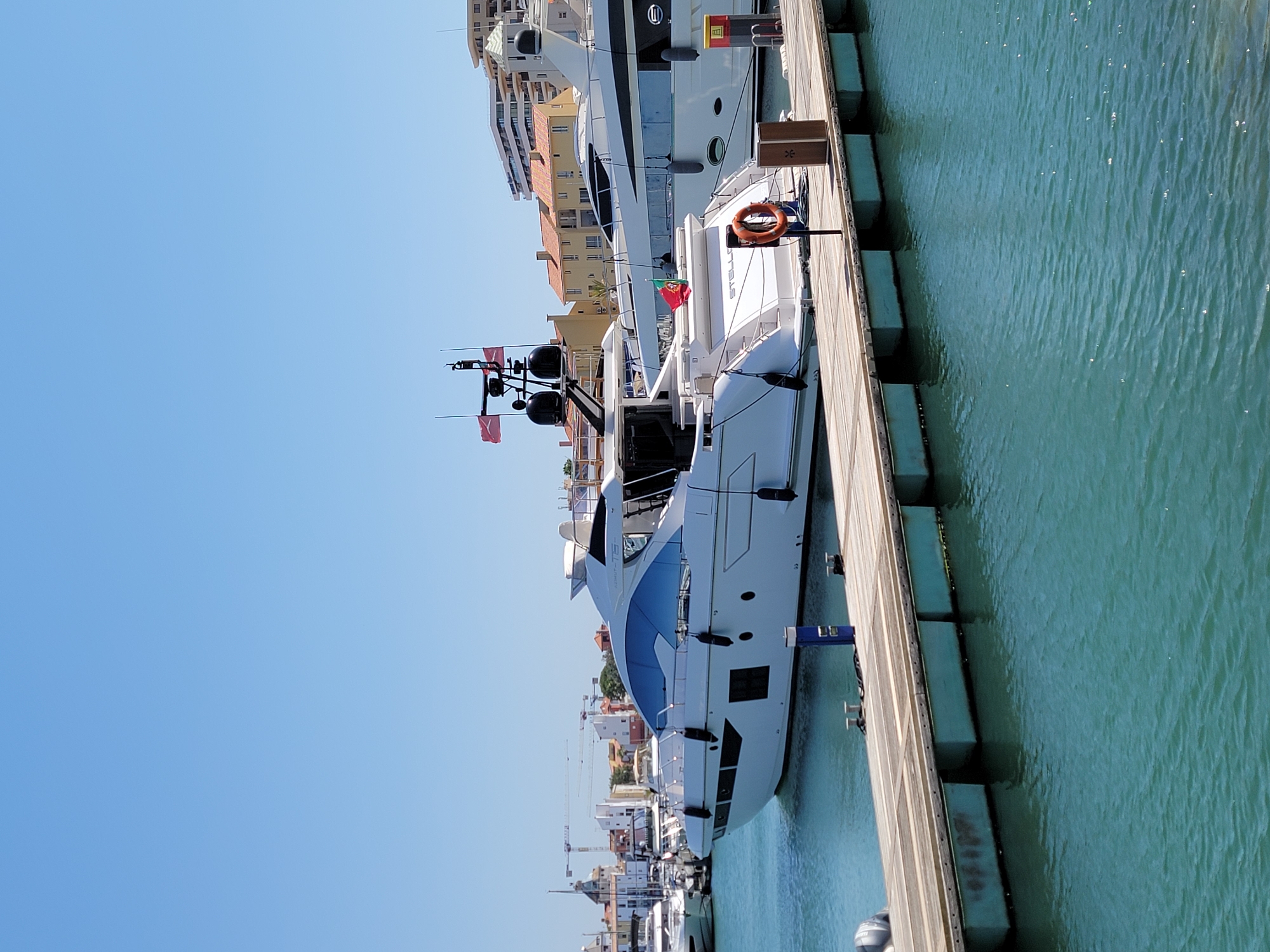 This image shows a marina in Quarteira, Portugal, on a clear, sunny day. The primary focus is a large, white luxury yacht docked at a wooden pier. The yacht has a sleek, modern design with dark tinted windows and several fenders along its hull. Another, larger yacht is visible behind it, also docked. In the background, there are buildings with terracotta roofs, typical of Portuguese architecture, and some construction cranes can be seen. The water in the marina is a clear turquoise color. There are no people visible in the image.