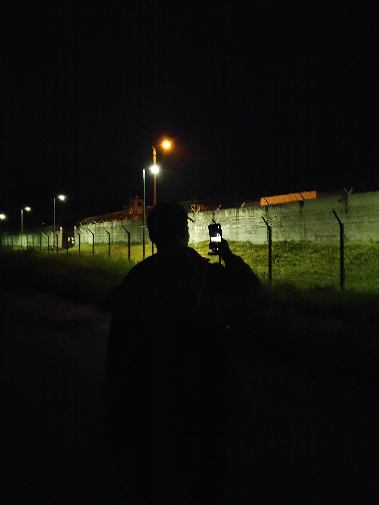 This image depicts a nighttime scene in the city of Chaikovsky, Russia. A silhouetted individual, seen from behind, occupies the lower central portion of the frame. This person's right arm is raised, holding a smartphone vertically, with its illuminated screen facing the viewer. To the left, a series of streetlights cast bright white and warm yellow light, illuminating a section of overgrown grass along a dark path. To the right of the person, a tall, light-colored concrete or stone wall topped with barbed wire or razor wire runs horizontally across the midground. A metal fence with pointed posts stands in front of this wall, with green grass visible between the fence and the wall. Further in the background, along the wall, a structure resembling a watchtower or a building within a secure facility is visible. The sky is dark, indicating night, and the scene is primarily lit by artificial streetlights.
