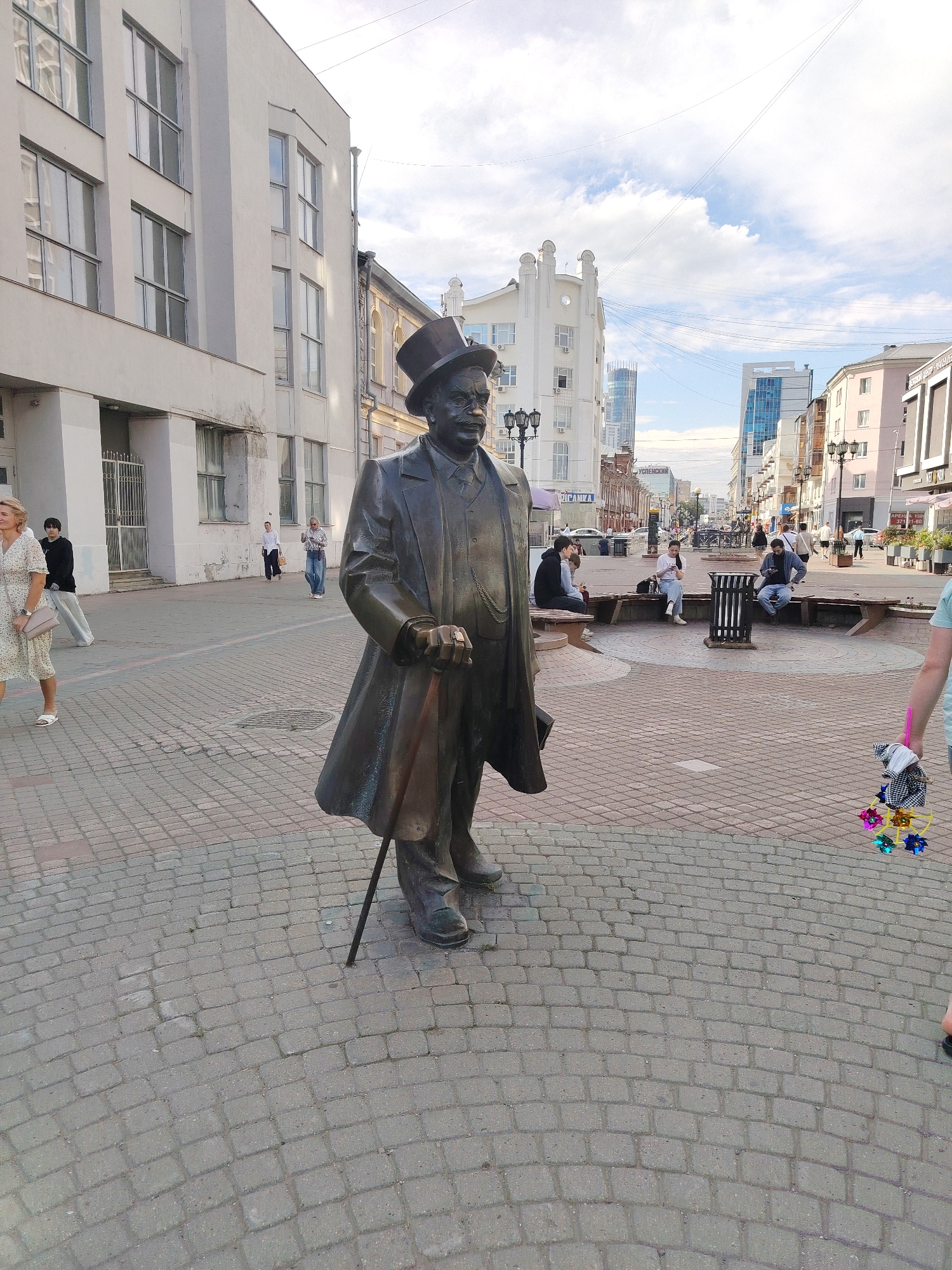 The image captures a street scene in Yekaterinburg, Russia, focusing on a bronze statue of a man in a top hat and coat, holding a cane. The statue stands on a paved plaza made of interlocking bricks. In the background, several buildings are visible, including a white, multi-story structure with large windows on the left, and other buildings with a mix of architectural styles lining a street further down. Several people are present in the scene: a woman in a light-colored dress walks on the left, a person in a black hoodie and light-colored pants stands near the building on the left, and a few individuals are seated on benches in the middle ground of the plaza. A person's arm holding colorful small decorative bows is visible in the lower right corner of the frame. The sky above is partly cloudy. The overall setting appears to be a public urban space.