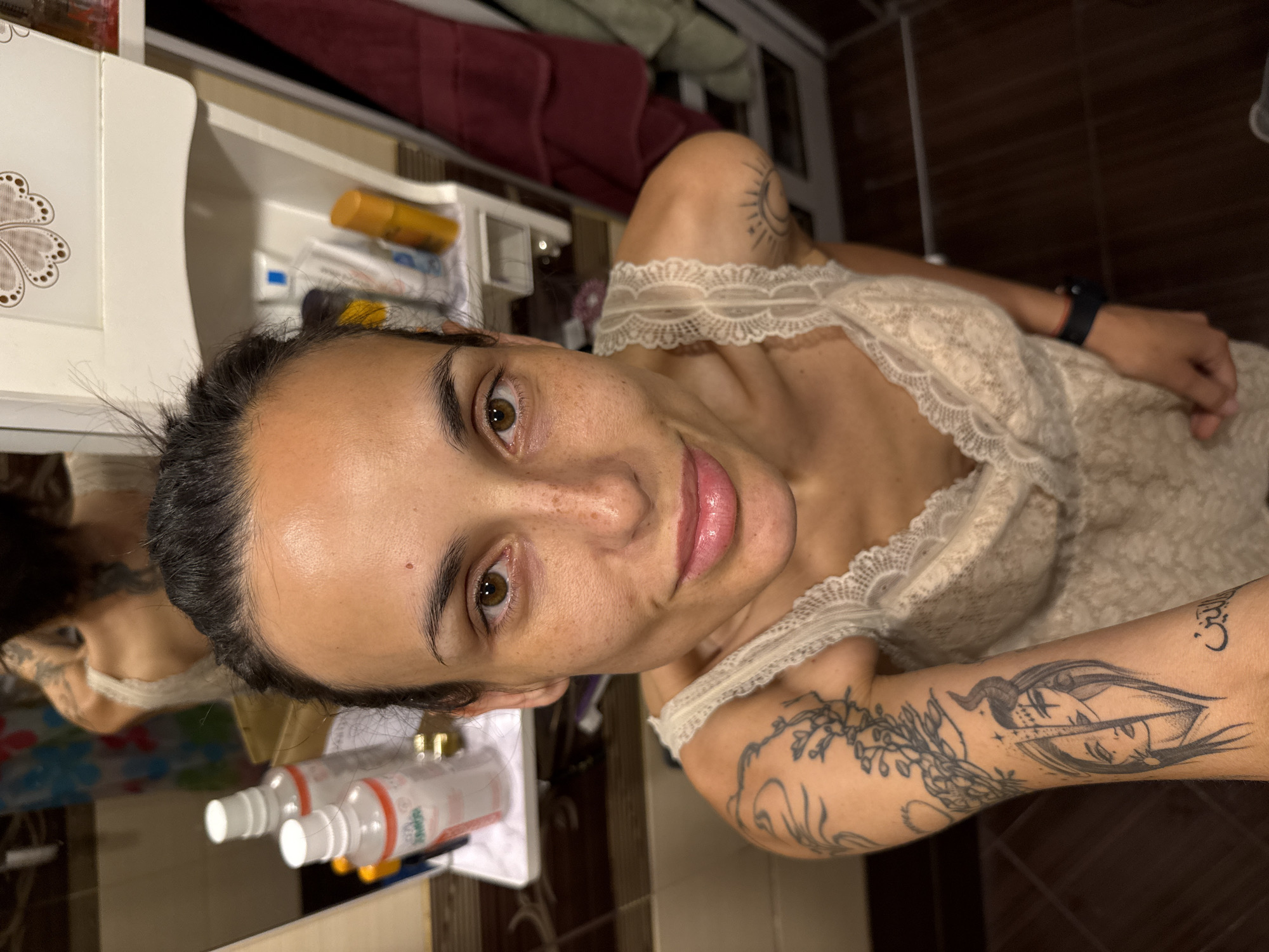 A selfie-style, close-up image captures a woman with olive skin, dark hair pulled back, and brown eyes. She is wearing a light-colored, lace-trimmed top. Visible on her arms are several tattoos, including what appears to be a woman's face and a plant-like design. The background suggests a bathroom setting, with a sink, toiletries, and brown-tiled walls. Two bottles with white caps and red writing sit on the edge of the sink in the foreground. The location or presence of the city of Pleven, Bulgaria, is not deducible from this image.