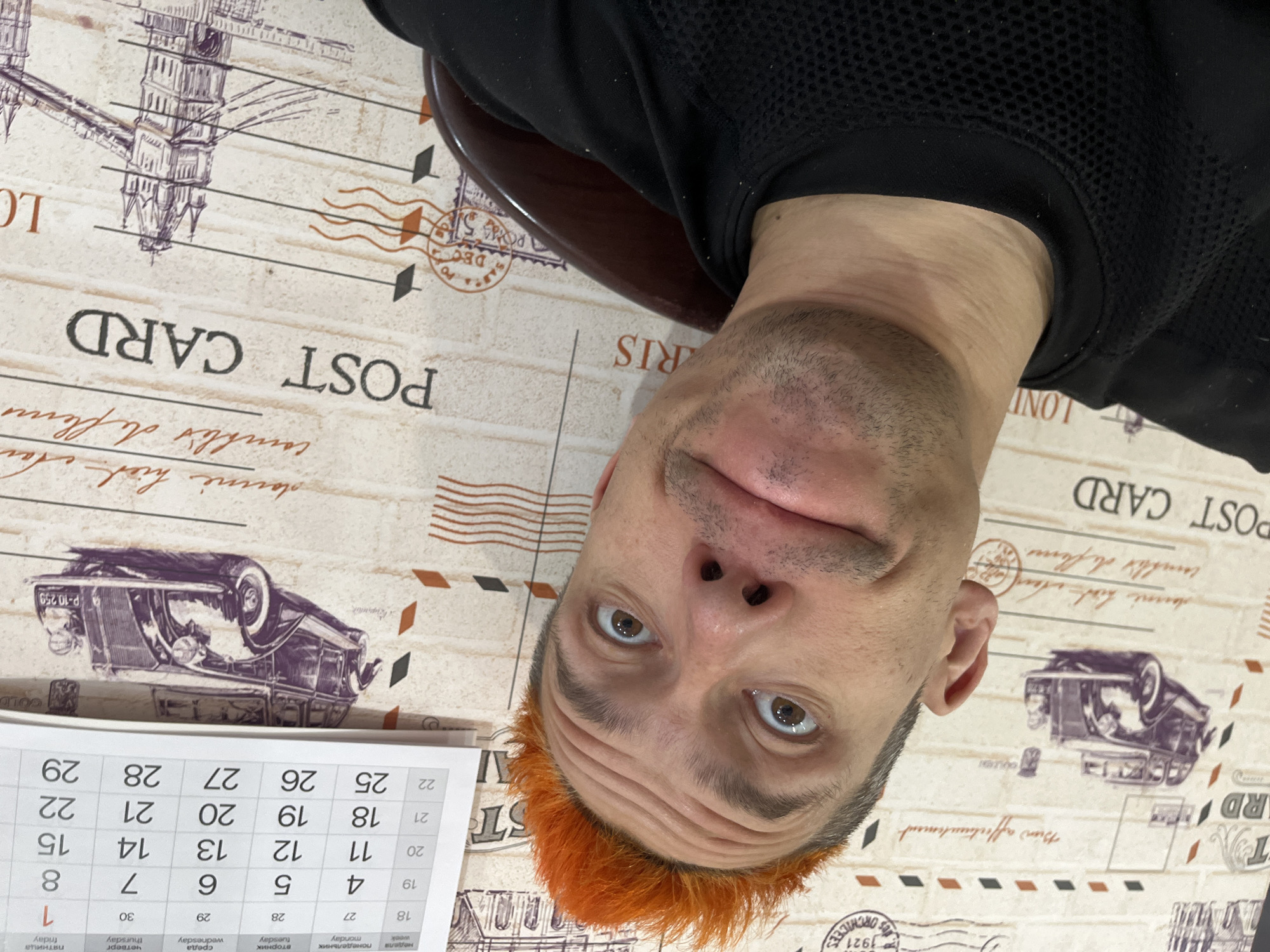 A man with orange hair is close to the camera, tilting his head back. He is wearing a black mesh shirt. He is positioned on a patterned surface that reads "POST CARD" and has images of cars and architectural designs. To the left of the man is a calendar. The calendar shows dates from 25-29 and 1-15 and 18-22, with Friday the first. The location appears to be indoors. The city of Chaikovsky, Russia cannot be determined from the image.