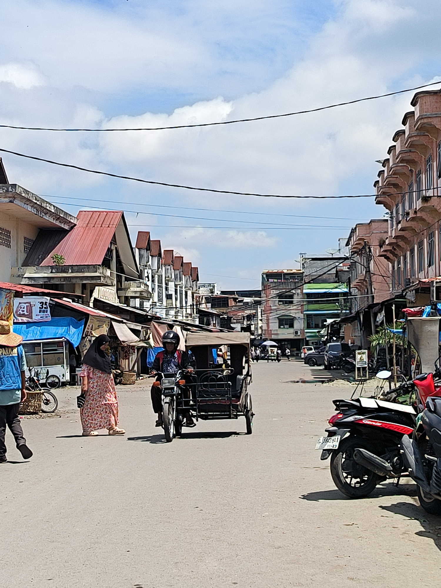 In Lhokseumawe, Indonesia, a street scene unfolds under a bright, cloudy sky. The road is paved and appears to be a main thoroughfare in a bustling area, lined with buildings of varying architectural styles and heights. On the left, a person wearing a blue vest and a straw hat walks away from the camera. Next to them, a woman in a floral dress and hijab stands, facing the viewer. In the center of the frame, a three-wheeled vehicle, likely a type of motorcycle taxi, is moving forward. The driver, wearing a helmet, is at the controls, and a passenger is seated behind them. The vehicle's canvas roof is extended.
Further down the street, other vehicles, including motorcycles and cars, are visible. Various stalls and shops line the sides of the road, some covered with tarps or awnings, suggesting a market or commercial district. Power lines crisscross overhead. The buildings exhibit a mix of older colonial-style architecture with balconies and more modern, functional designs. The overall impression is one of everyday life and commerce in a Southeast Asian city.