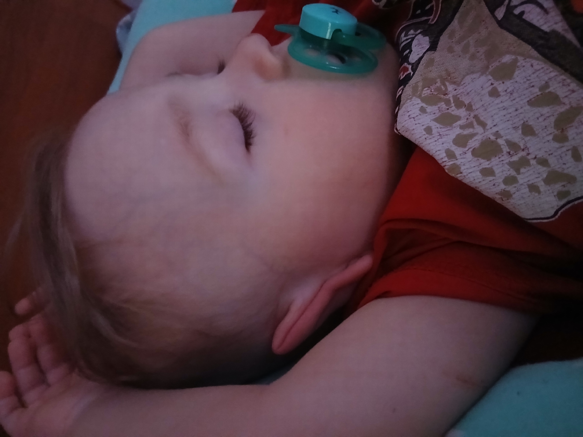 A close-up image depicts an infant sleeping on its right side, facing towards the right of the frame. The infant has light-toned skin and light-colored hair, partially visible on the head. Eyelids are closed, with visible eyelashes. A teal-colored pacifier is inserted into the infant's mouth. The infant is wearing a red garment, possibly a shirt or pajama top, with a portion of patterned fabric visible beneath, featuring light irregular shapes on a white background. The infant's left arm is raised, with the hand positioned near the top of the head. A light blue fabric object, possibly a blanket or pillow, is visible underneath the infant's neck and shoulder. The background is dimly lit, with a dark brown surface visible in the upper right. The image provides no visual information to confirm the specified location of Chaikovsky, Russia.