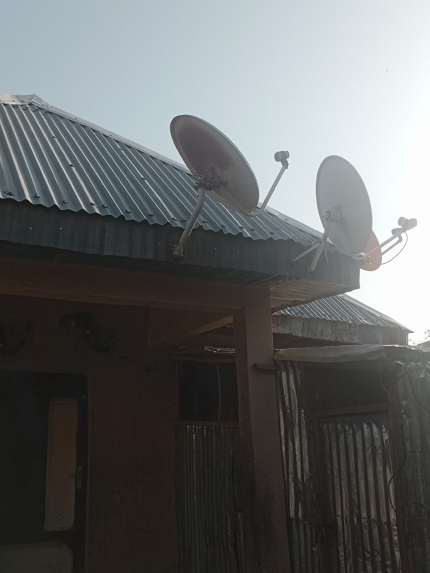 The image depicts the exterior of a residential building with a corrugated metal roof in Jalingo, Nigeria. Two satellite dishes are mounted on the roof, angled towards the sky. The building appears to be constructed with a combination of materials, including what looks like mud or plastered walls, and corrugated metal sheeting. A doorway with a vertical window is visible on the left side of the building, and further to the right, a section of the building is covered with corrugated metal. The overall impression is of a typical urban dwelling in Nigeria.
