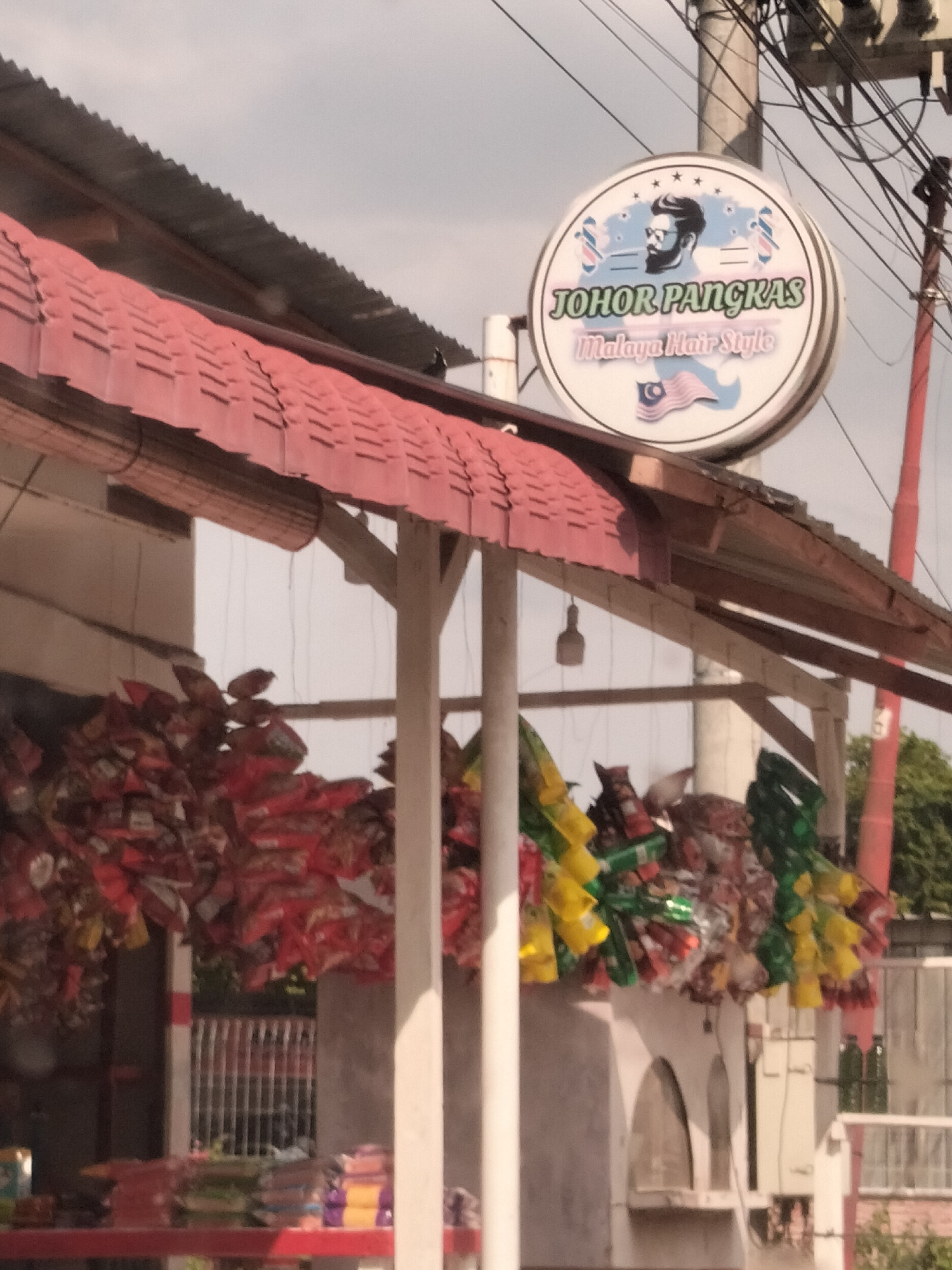 The image depicts a storefront under a red-tiled awning, seemingly a small business located in Titibaro, Indonesia. Strings of multi-colored snack bags hang beneath the awning. A sign indicates a barber shop called "Johor Pangkas Malaya Hair Style." A utility pole with wires is visible in the background. The lighting suggests an overcast day.