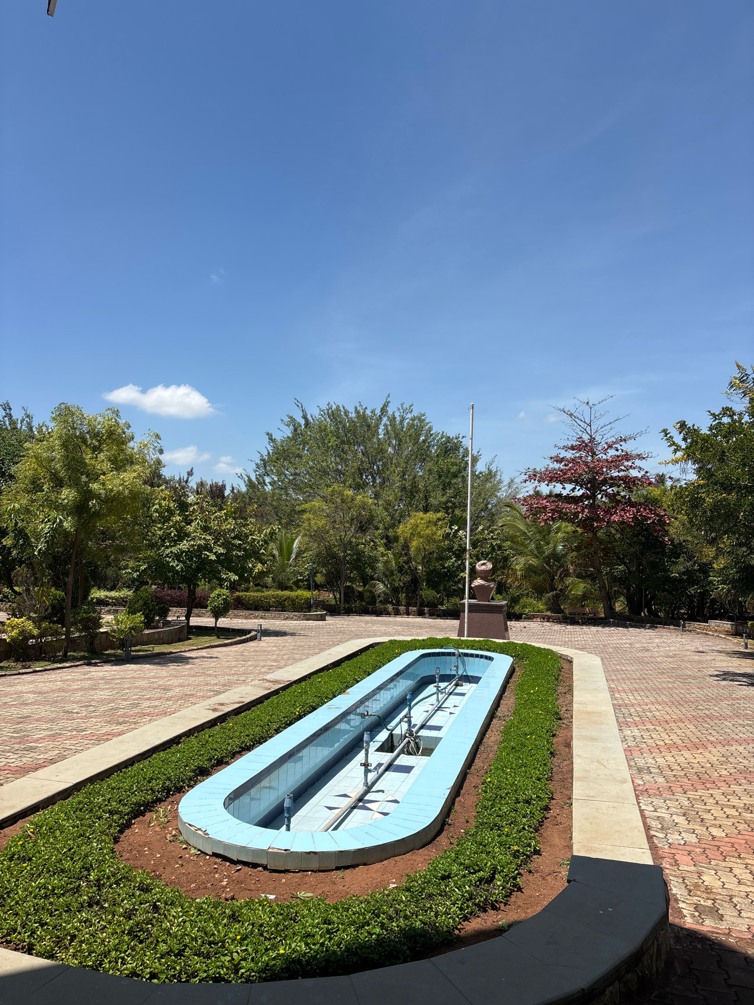 This is a medium-shot, eye-level, outdoor photograph captured on a sunny day in the city of Jhangatihala, India. The foreground features a long, rectangular, light blue fountain that appears to be empty. The fountain is surrounded by a manicured hedge of dark green, leafy plants. Beyond the fountain, a pathway paved with red and beige interlocking bricks curves towards the background. To the left of the pathway, there are more trees and bushes. In the middle ground, a flagpole stands to the left of a reddish-brown statue on a dark pedestal. Behind the statue, a tree with vibrant red leaves is prominent. The background is filled with a dense arrangement of various green trees of different sizes and textures, under a clear blue sky with a few wispy white clouds. The overall scene depicts a well-maintained public space or park.