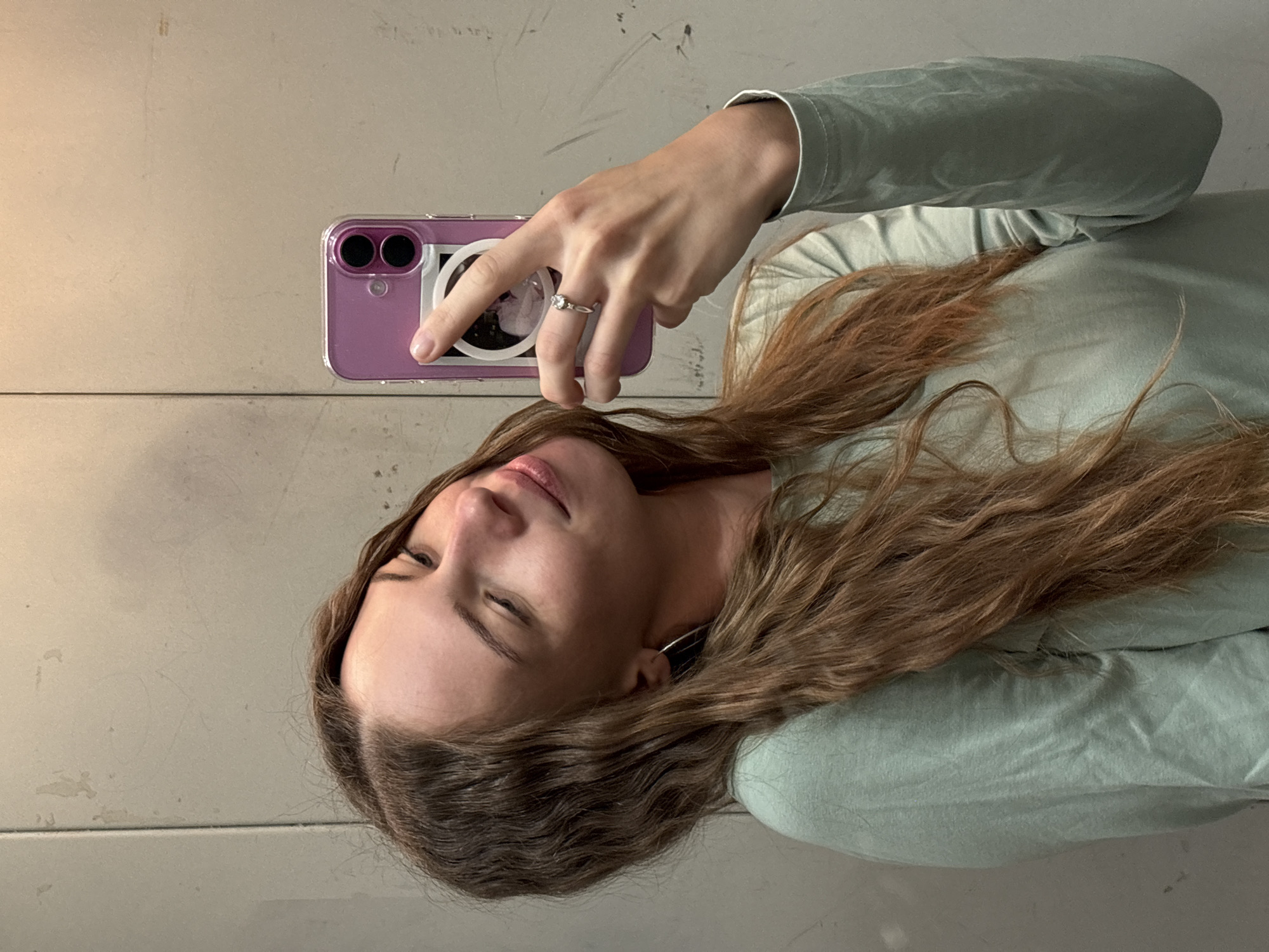 A young woman with long, wavy brown hair is taking a selfie in a mirror. She is wearing a light green long-sleeved shirt and a silver ring on her left ring finger. Her phone, with a pink case, is held up to take the picture. The background appears to be a grey, textured wall. The location is not identifiable as Perm, Russia.