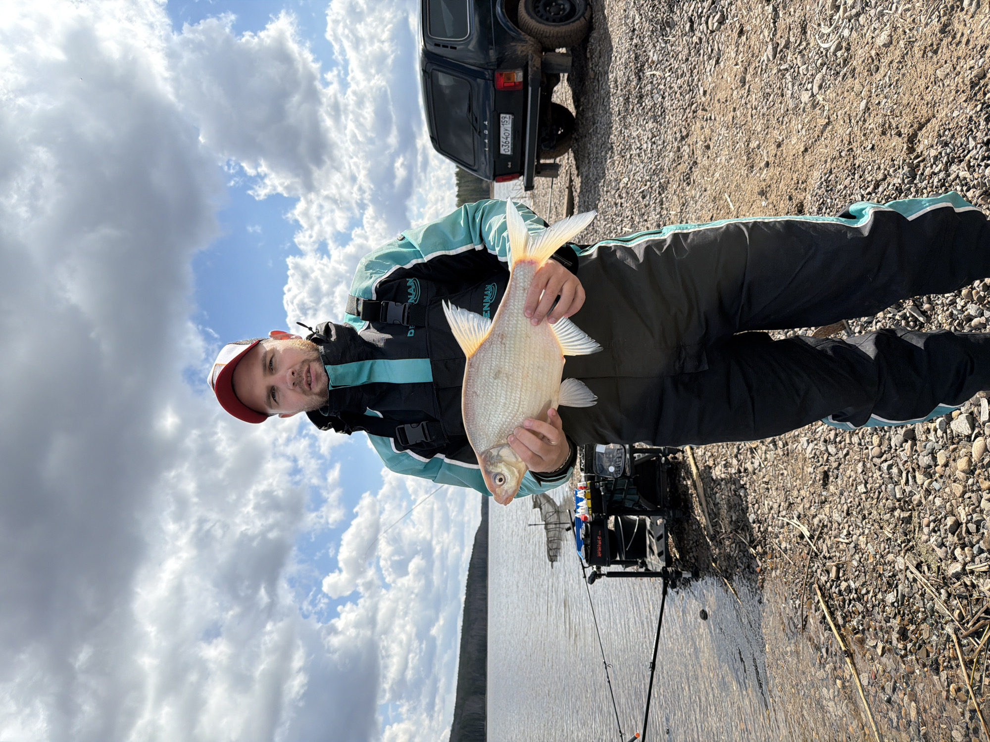 The image, correctly oriented, captures a man holding a large fish by a body of water in Chaikovsky, Russia. The man, appearing to be in his 30s or 40s with a light beard, wears a red and white cap and a two-tone black and light blue fishing suit with "DRENNAN" visible on the chest. He presents a large, light-colored fish, possibly a bream, with a slight smirk of satisfaction.
The setting is a natural, outdoor environment, likely a riverside or lakeside. The man stands on a gravelly and rocky bank. In the background, to his right, a dark Lada Niva SUV is partially visible, parked on the similar terrain. The vehicle's license plate, "0364OY 159," is clearly discernible. Near the water's edge, specialized fishing gear, including multiple fishing rods and what appears to be a fishing chair or platform with various accessories, indicates an organized fishing trip.
The body of water extends into the distance, with a dark, tree-lined or hilly horizon on the opposite bank. The sky is dominated by a mixture of large, fluffy white and grey clouds, suggesting an overcast but bright daytime. The "159" region code on the license plate confirms the Perm Krai location, aligning with Chaikovsky. The image conveys a sense of quiet accomplishment and enjoyment of nature.