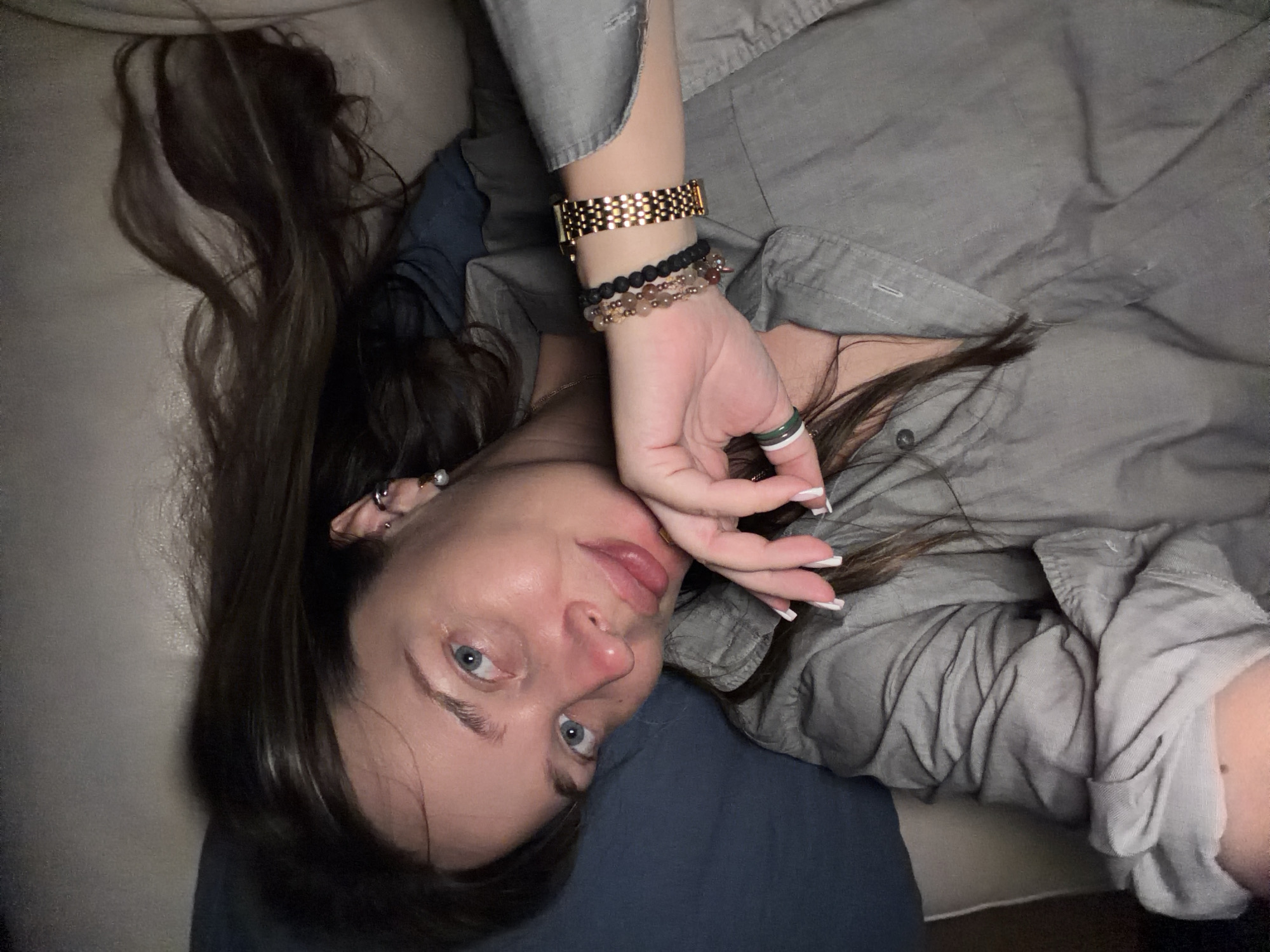 A woman is lying on a couch, looking at the camera. She has long, dark brown hair and blue eyes. She is wearing a gray button-up shirt and several bracelets on her left wrist. Her hand is resting on her chin. The background is a beige couch and a dark blue pillow. The image is taken indoors.