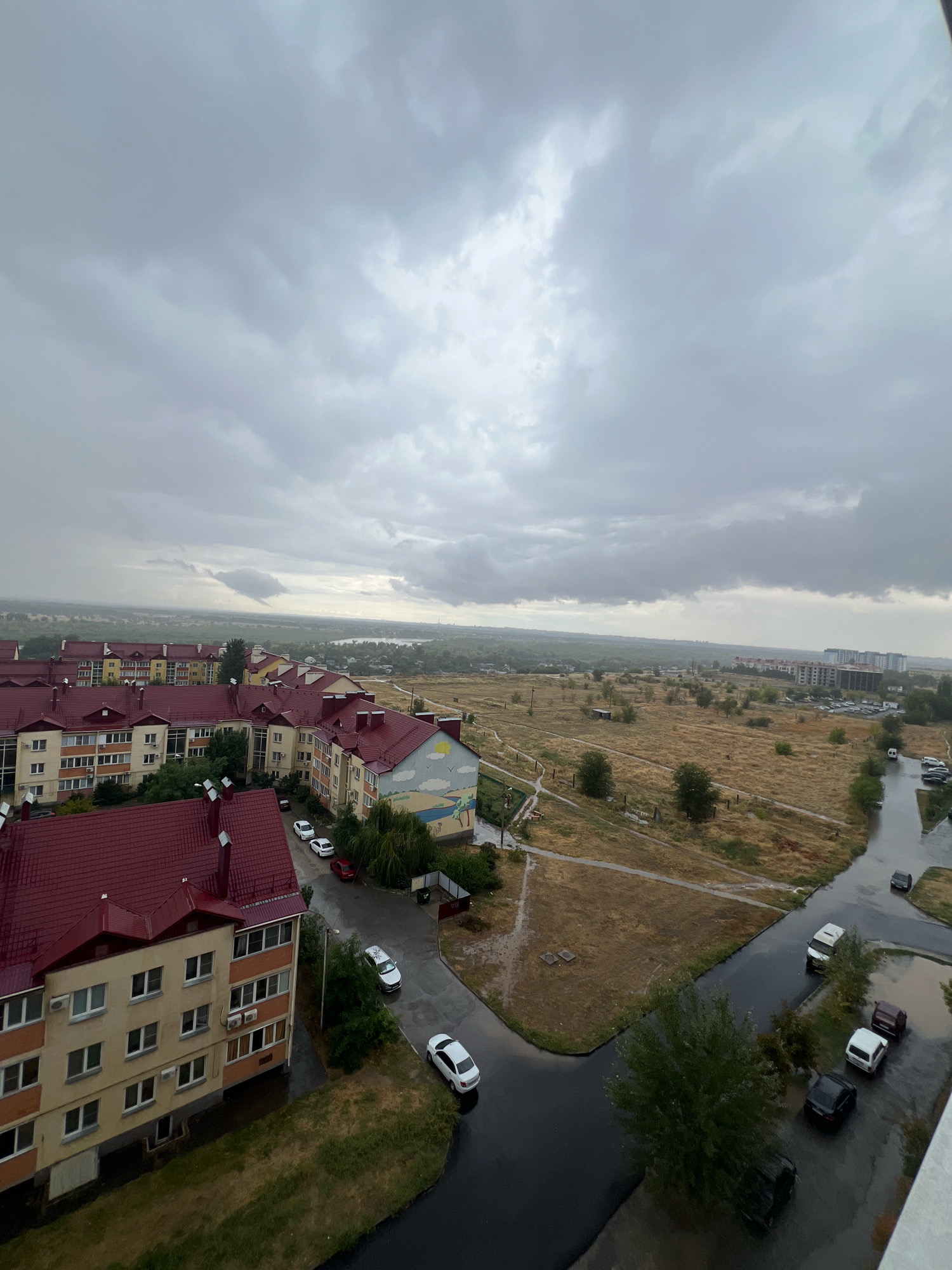 The image depicts an aerial view of a residential area in Volzhsky, Russia, under a cloudy sky with signs of recent rain. The foreground features several apartment buildings with distinctive red tiled roofs. These buildings are multi-story, with cream-colored facades and numerous windows. Some of the buildings have murals on their sides, including one depicting a body of water with birds and a sun.
A wet asphalt road winds through the scene, with several cars parked or driving on it. The road is reflecting the overcast sky and appears to have puddles, indicating ongoing or recently concluded rainfall. Surrounding the buildings are patches of green grass and dry, yellowish vegetation. Beyond the immediate residential area, the landscape transitions to a more open, field-like terrain with scattered trees and some smaller structures. In the distance, more apartment buildings are visible, along with a hazy horizon suggesting a larger urban expanse or natural landscape. The overall atmosphere is somber, consistent with overcast weather.