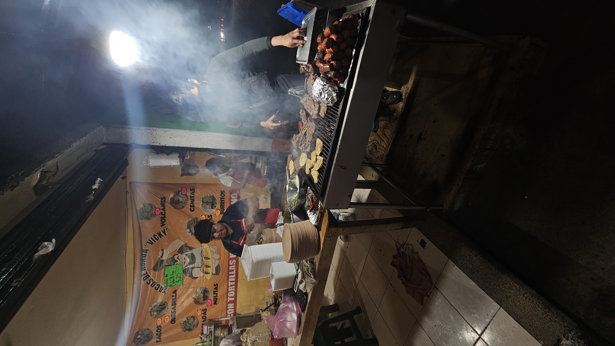 The image shows a street food stall in San Bernardino Tlaxcalancingo, Mexico, selling tacos, quesadillas, and other Mexican food. A cook wearing an apron is preparing food, while another person appears to be operating the grill. The stall is decorated with a banner advertising the food, and there is smoke rising from the grill. The surrounding area seems to be a street corner with dark walls and tiled floors. Plates and food preparation items are visible on the counter, suggesting active food preparation.