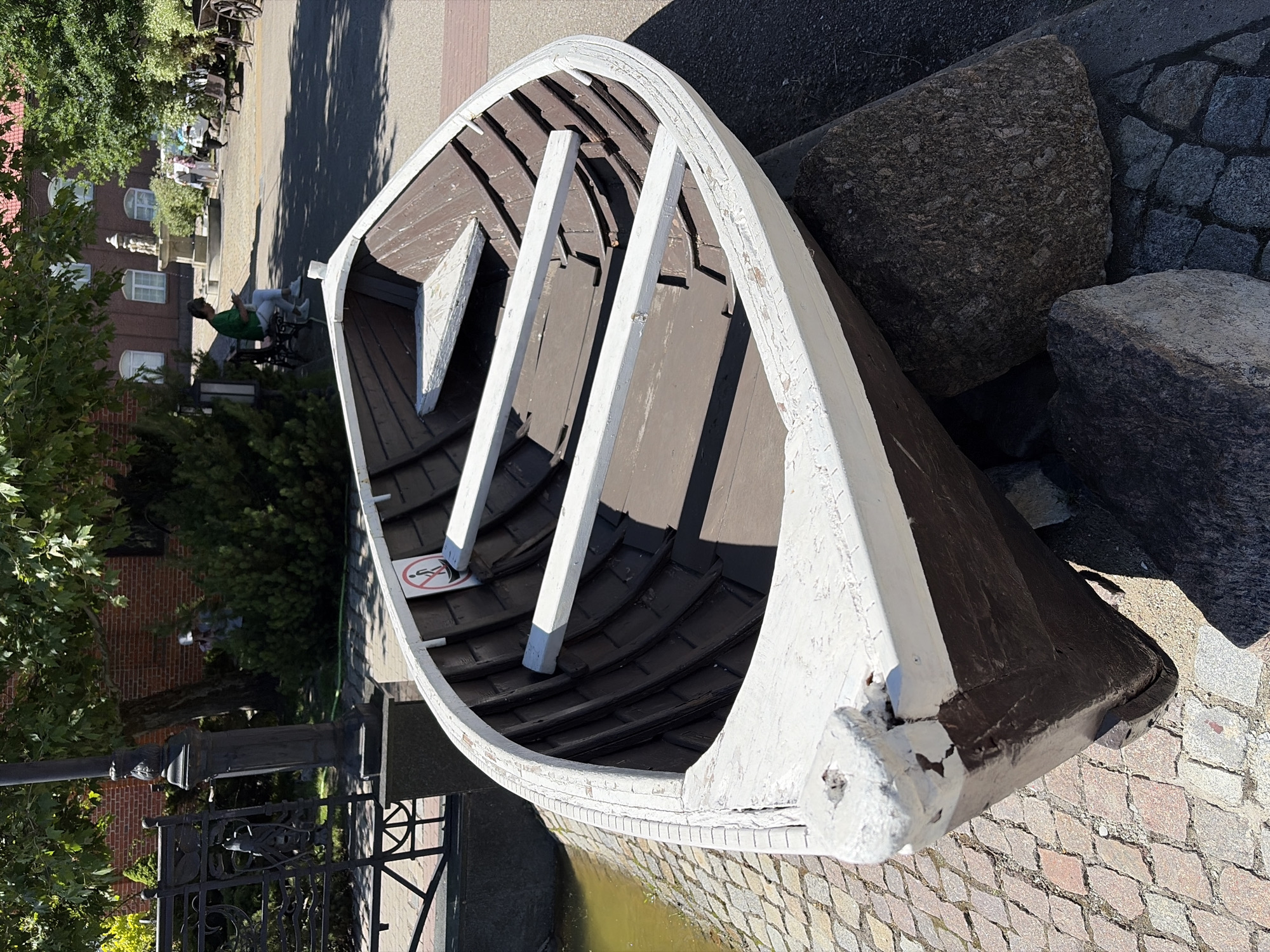 An old, brown and white wooden rowboat rests on cobblestones and large rocks. The boat is angled and partially supported by the rocks. Inside the boat, two wooden oars are visible, along with a small white seat at the front and a sign with a person in a circle with a line through it. Behind the boat, a cobblestone path leads to a paved area with trees and buildings. In the background, several people are present, including one sitting on a bench and another on a wheelchair. A lamppost and a decorative metal fence are also visible. The scene is outdoors on a sunny day.