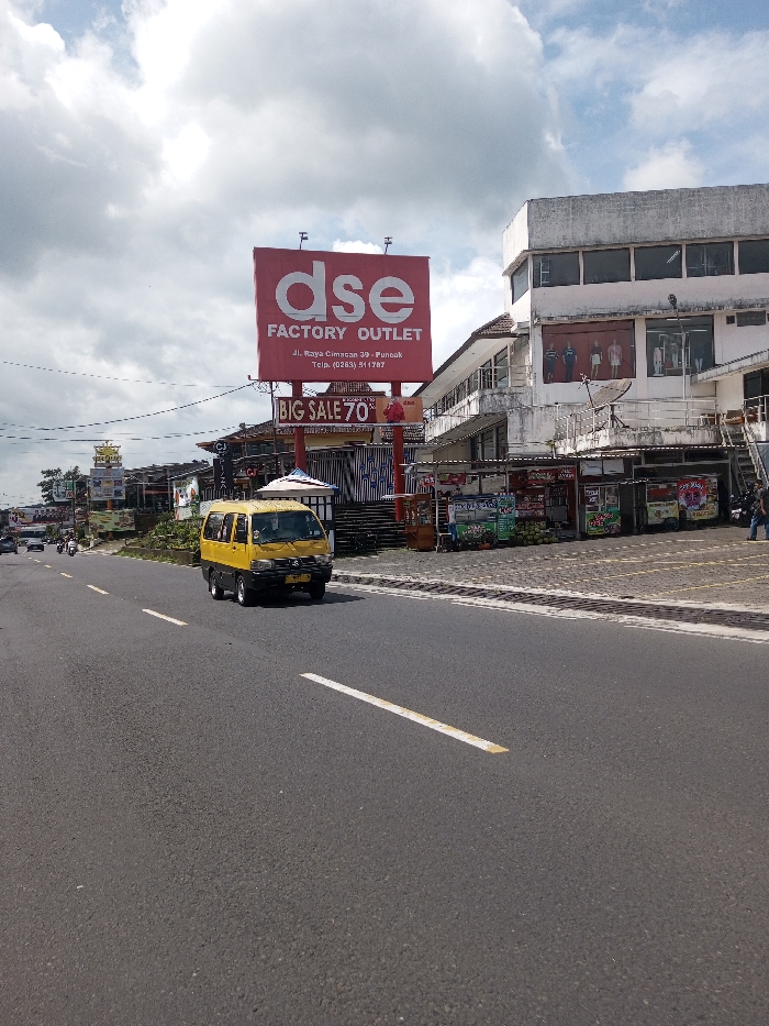 The image depicts a daytime street scene, identified as being in the city of Gubbisamamidi, India, under an overcast sky. A two-lane asphalt road with solid and dashed white markings extends into the distance. A yellow van, traveling away from the viewer, occupies the right lane in the foreground. Multiple vehicles, including motorcycles, are visible further down the road on the left.
On the right side of the road, a prominent red billboard displays "dse FACTORY OUTLET" in white lettering, with the address "Jl. Raya Cimacan 39 Puncak" and telephone number "(0263) 511787" below it. A smaller red sign beneath the main billboard advertises "BIG SALE 70%." Adjacent to the billboard stands a multi-story commercial building with a light-colored, weathered exterior. Its upper floor windows showcase clothing on mannequins, and a satellite dish is attached to the facade. The ground level features several vendor stalls, one displaying green coconuts. Multiple individuals are present on the sidewalk in front of these stalls. Additional commercial structures, utility poles, and signage line the street in the background.