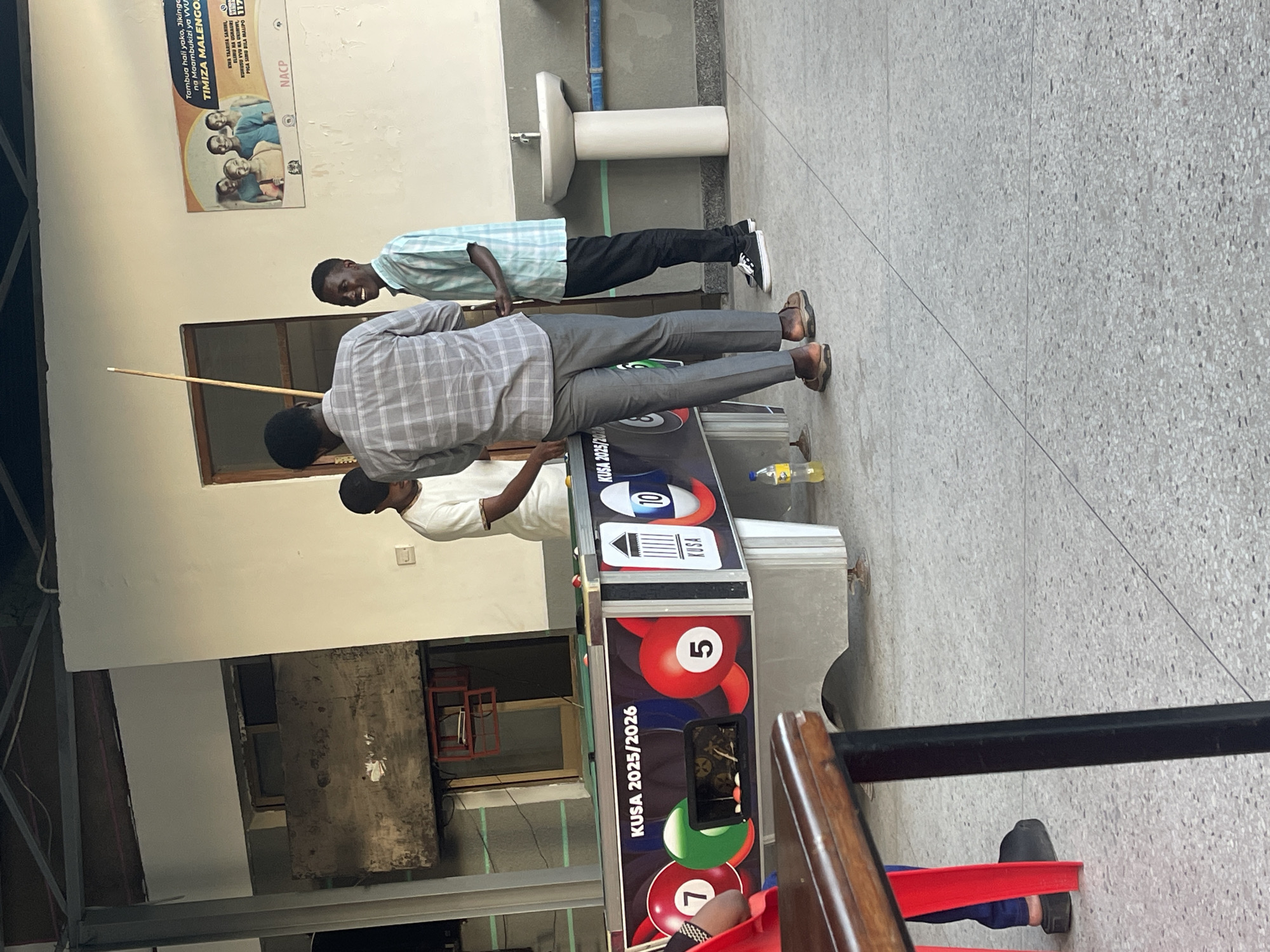 The image, taken in Dar es-Salaam, Tanzania, depicts three men playing or observing a game of pool in a casual, semi-outdoor setting. In the foreground, a man in a grey plaid shirt is bent over a black and silver pool table, holding a cue stick, focusing on the game. Another man in a white t-shirt stands close by, observing the play. Further back, a third man wearing a light blue patterned shirt and dark pants stands, looking towards the table with a broad, cheerful smile, indicating a relaxed atmosphere.
The pool table features colorful billiard balls and has "KUSA 2025/2026" and "KUSA" printed on its side, along with numbers like 5, 7, and 10. A plastic bottle rests on the table. The background includes a light-colored wall with a white wall-mounted sink and faucet. Above the sink, a poster written in Kiswahili is displayed.
The poster conveys public health messages: "Tambua hali yako, Jikinge na Maambukizi ya VVU" (Know your status, Protect yourself from HIV infection) and "TIMIZA MALENGO" (Achieve your goals). It also mentions "KUHUSU VVU NA UKIMWI" (About HIV and AIDS) and provides a toll-free number: "PIGA SIMU BILA MALIPO 117" (Call toll-free 117), along with the acronym "NACP." The floor is a light grey speckled terrazzo tile. The scene suggests a friendly social gathering.