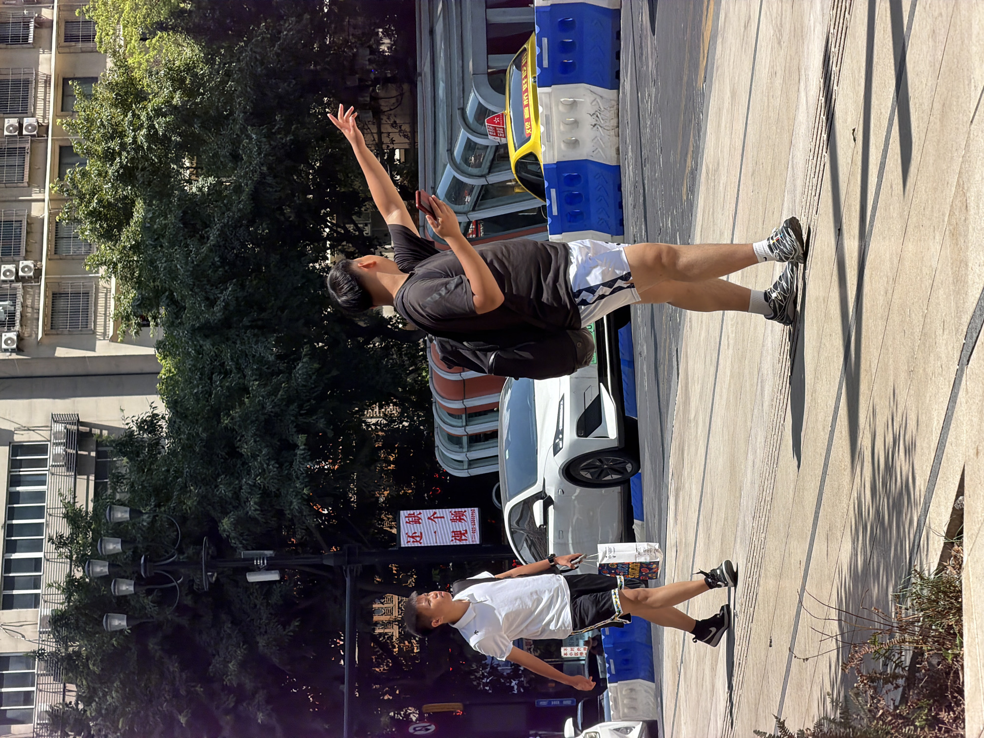 A medium shot captures two young men walking on a sunlit city sidewalk in Yuzhong District, China. The man on the left, wearing a white polo shirt and black shorts, carries a shopping bag in his left hand and walks with his head turned to the left. The man on the right, seen from the side, wears a black t-shirt, white shorts with a geometric pattern, and athletic shoes. He has a black backpack on and holds a smartphone in his right hand while extending his left arm and hand upwards, as if hailing a taxi or gesturing. A white car is visible behind the man on the left, and a yellow taxi is behind the man on the right, partially obscured by blue and white construction barriers. In the background, a building with multiple windows and air conditioning units stands behind a line of trees, with a street lamp on the left side of the frame. The sidewalk is made of concrete with visible darker lines.